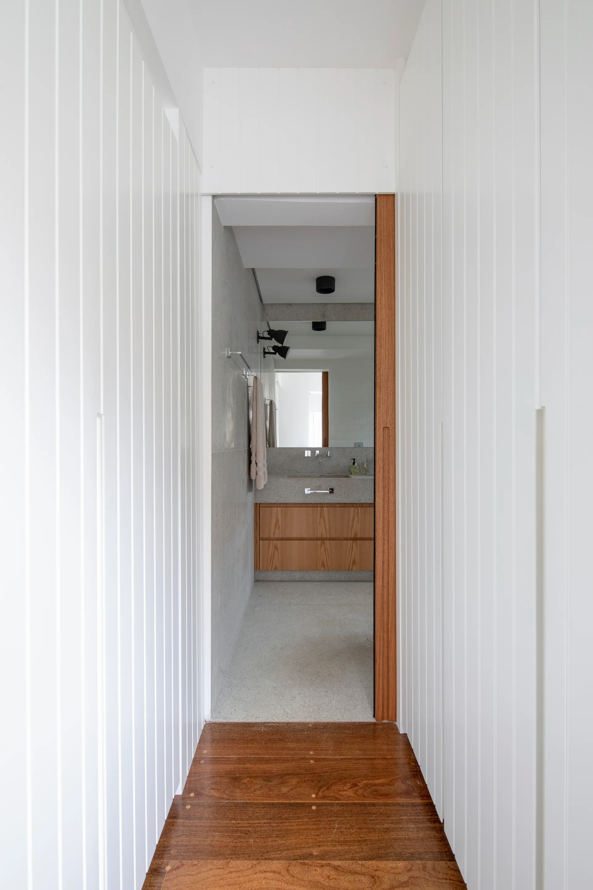 A view through a white vertically paneled hallway with wood flooring into a modern bathroom featuring a wood vanity and stone walls.