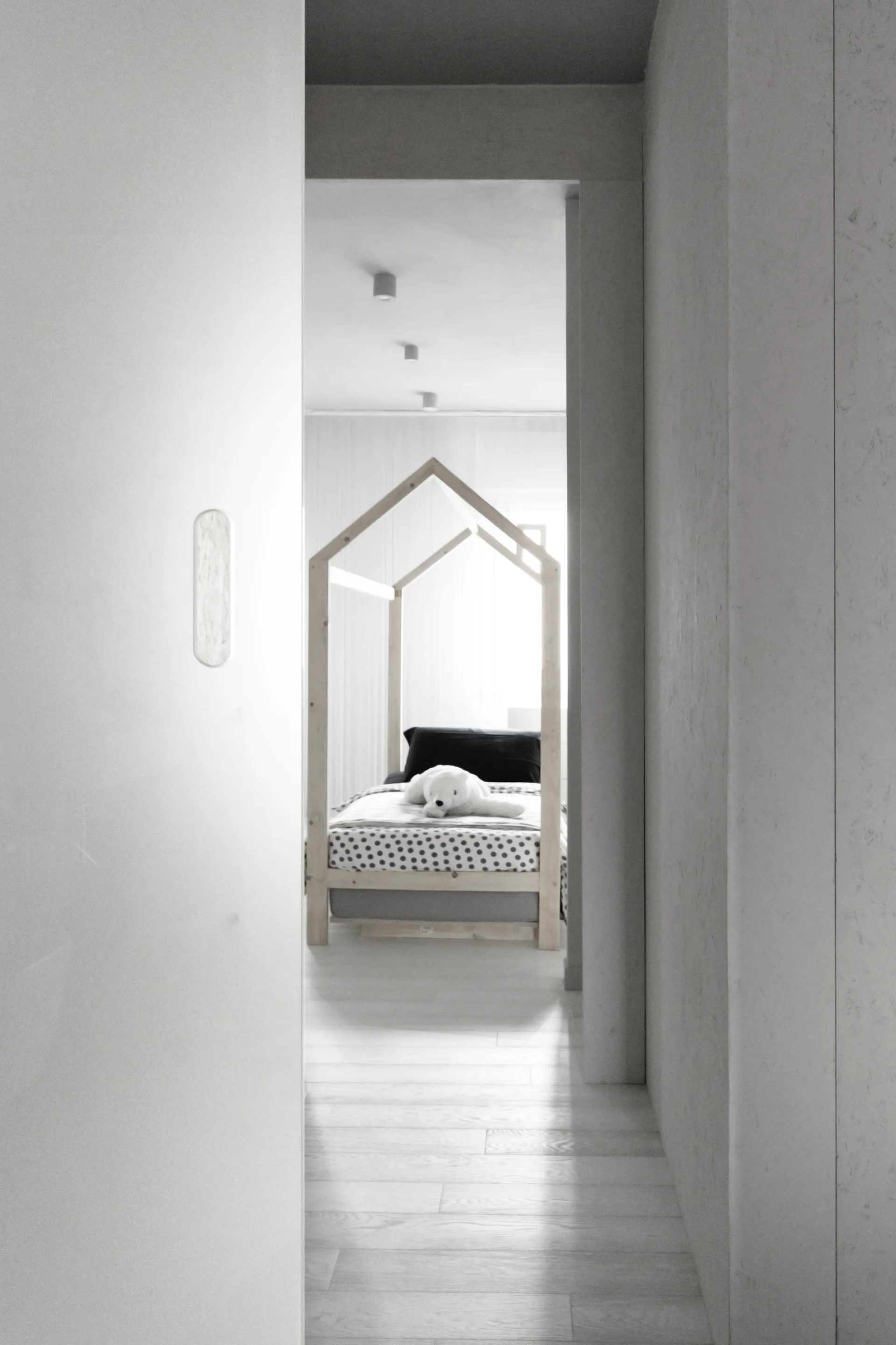 A view through an open doorway into a minimalist child's room featuring a wooden house-shaped bed frame with polka dot bedding.