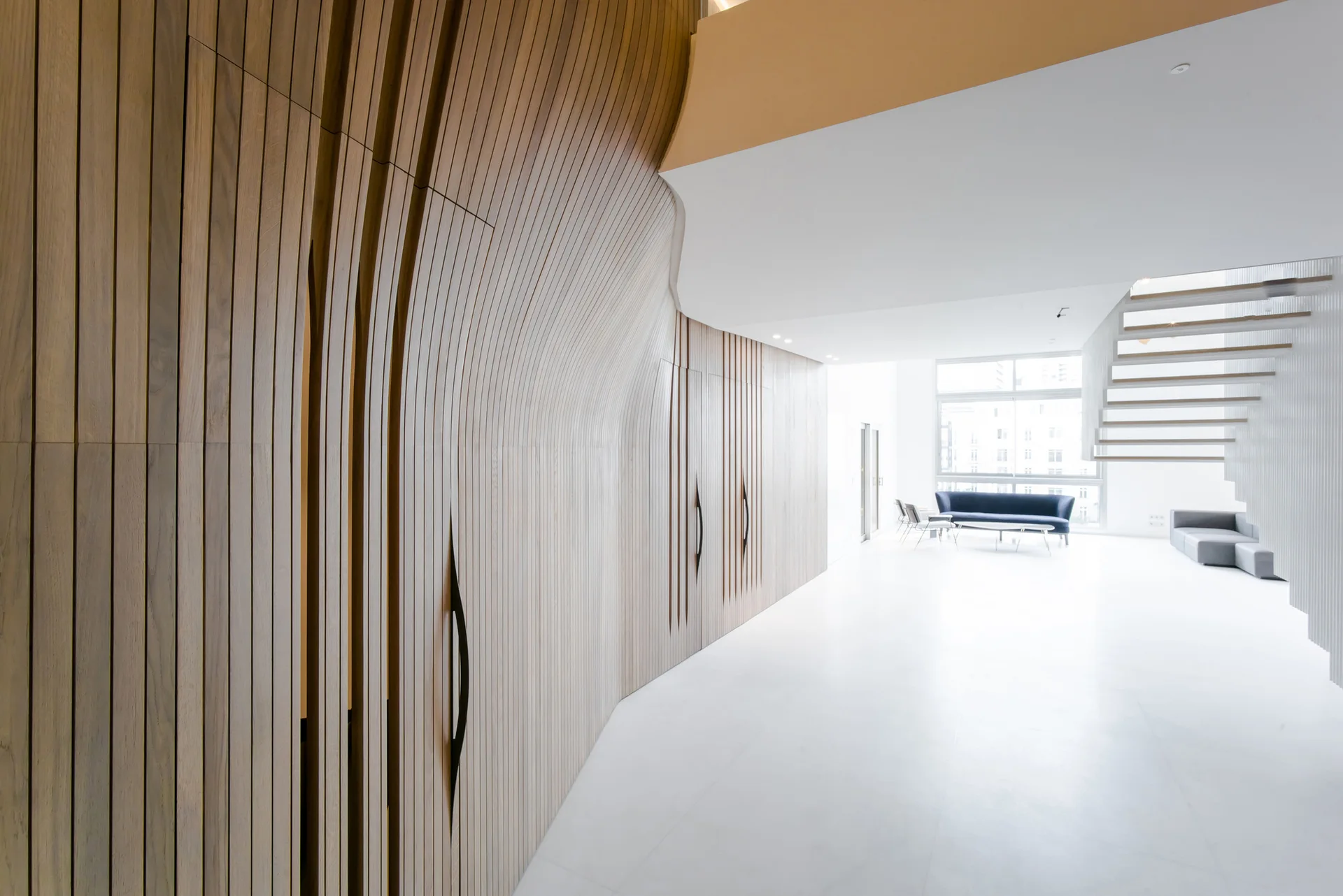 A minimalist white living room features a sweeping curved wood slat wall with integrated closets, a floating staircase, and distant blue seating.