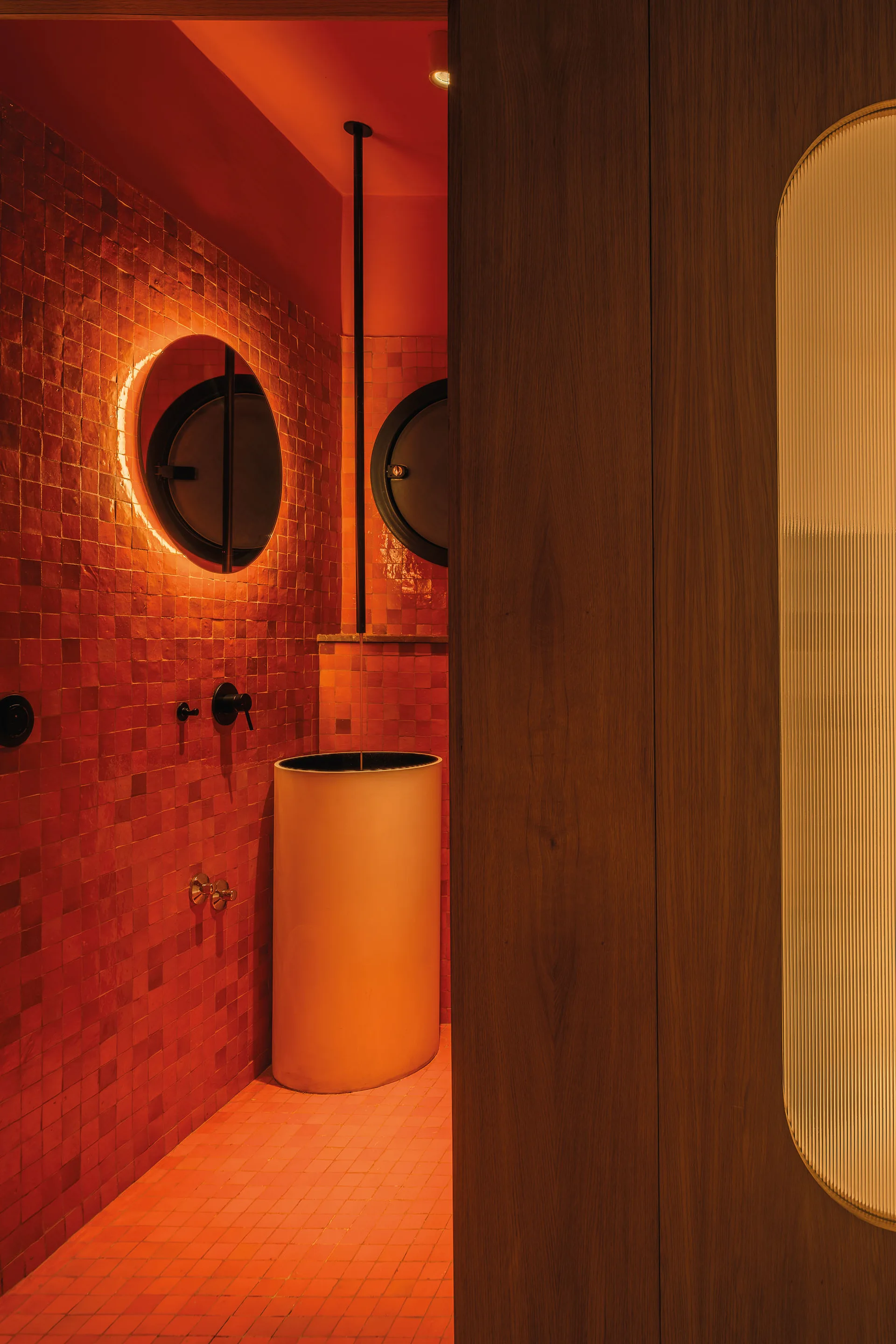 A small powder room with glossy red tiled walls, a tall white cylindrical pedestal sink, and a backlit circular mirror.