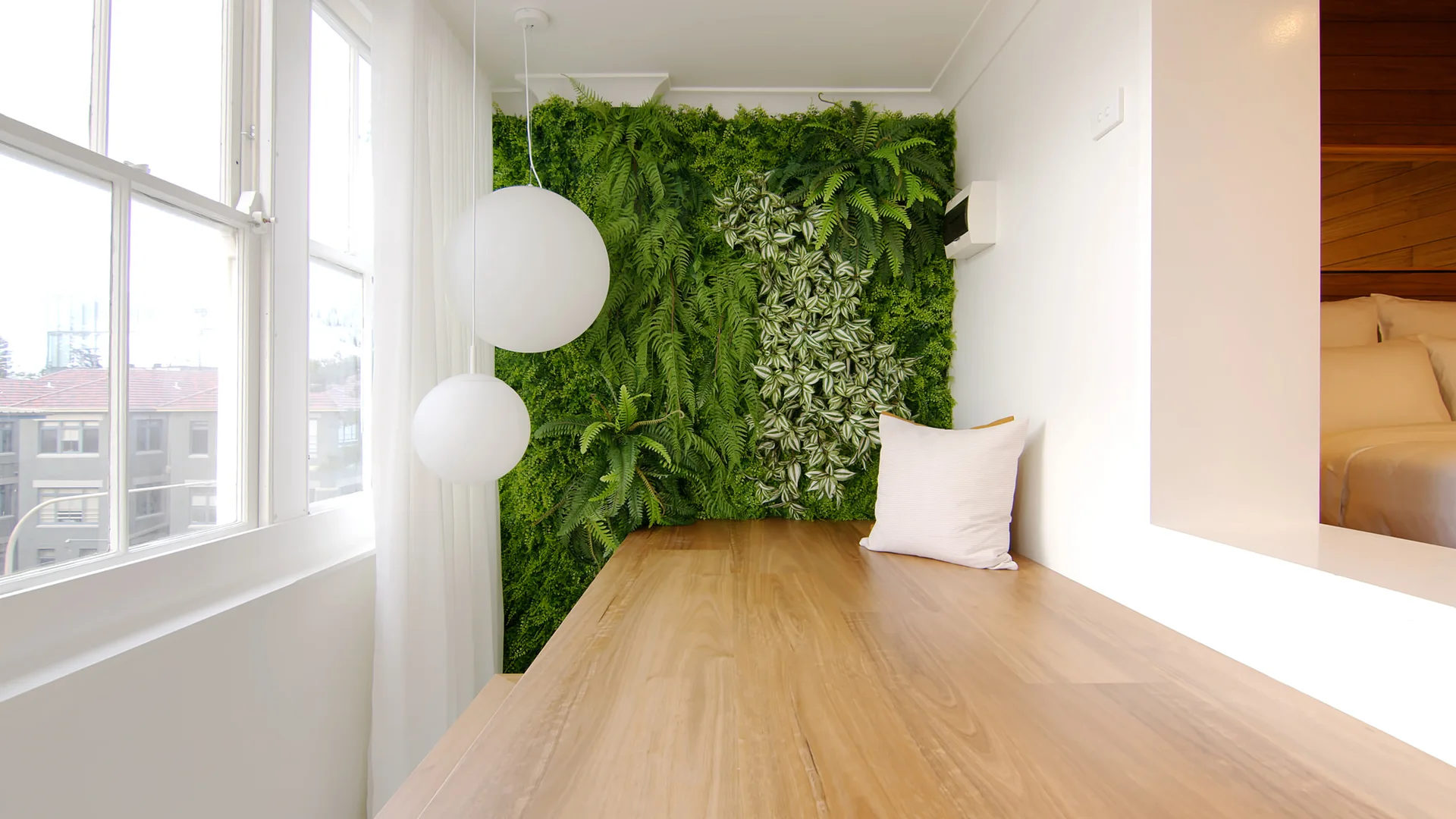 A minimalist bedroom alcove features a built-in wood platform, a vertical green plant wall, and two hanging white spherical pendant lights near a window.