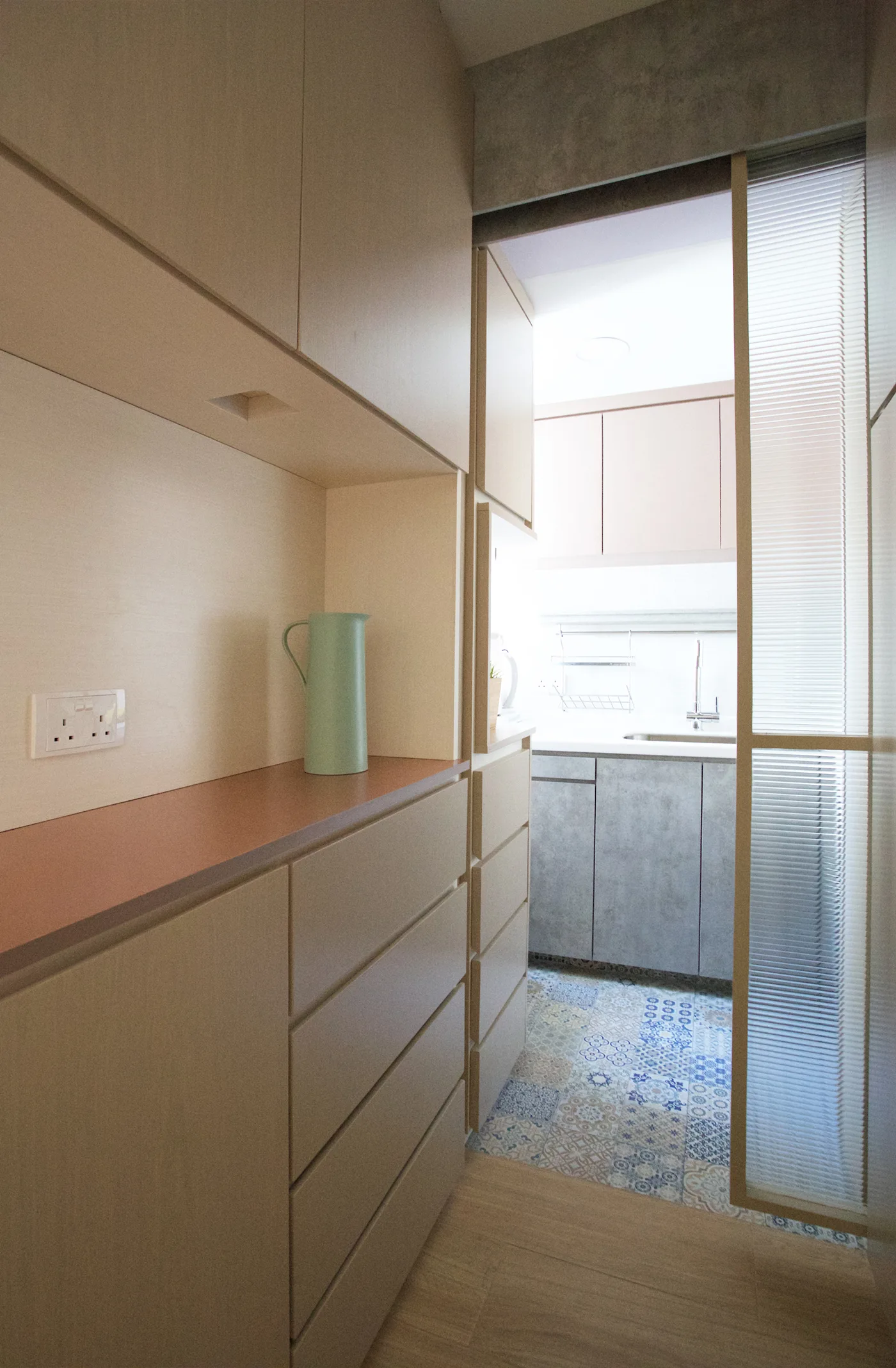 A narrow kitchen corridor features light wood built-in cabinets, a mint green pitcher, and a fluted glass sliding door opening to a patterned tile floor.