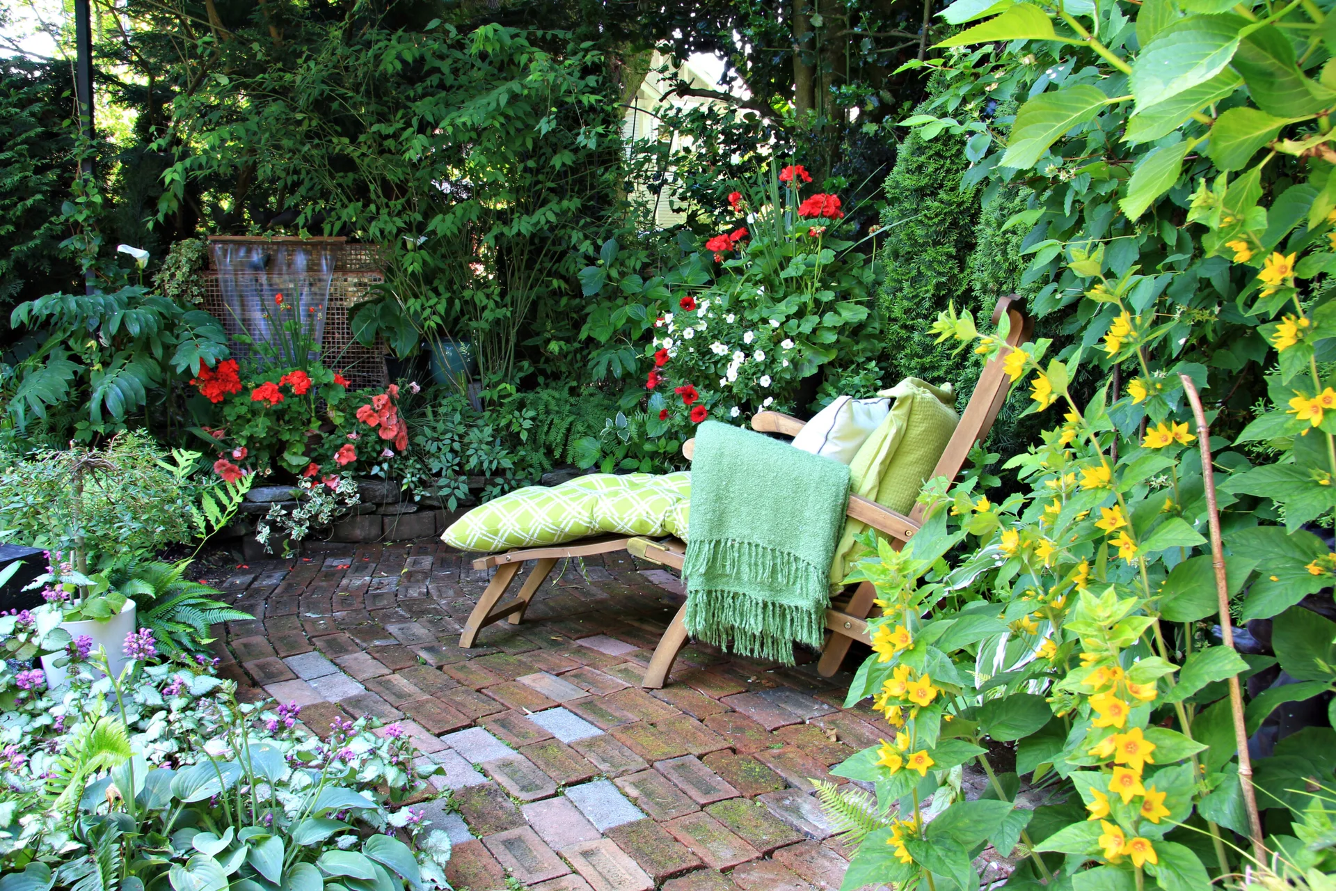 A wooden chaise lounge with green cushions sits on a weathered brick patio surrounded by dense, lush garden foliage and colorful flowers.