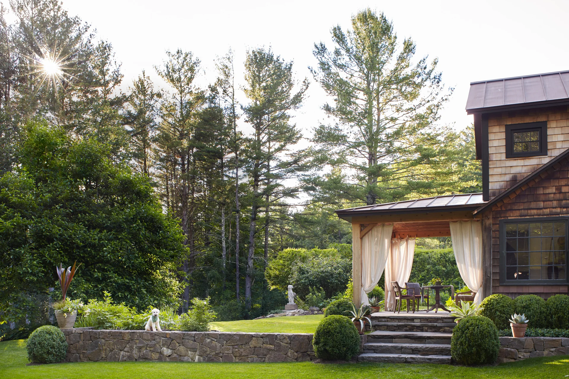 A covered rustic patio with light-colored curtains and stone steps is set next to a grassy lawn with a stacked stone retaining wall.