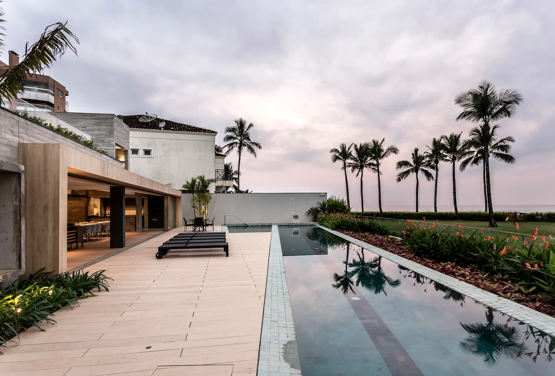 A long rectangular lap pool is flanked by a light wood deck with black lounge chairs and a row of palm trees overlooking the coast.