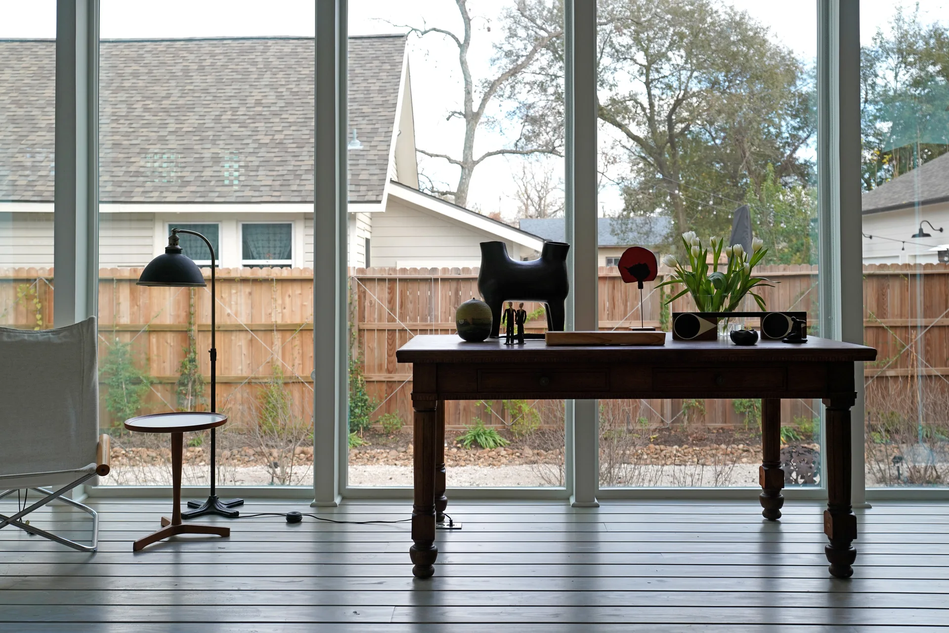 A dark wooden table with sculptural decor stands on a grey plank floor in front of floor-to-ceiling windows looking out to a wooden fence.