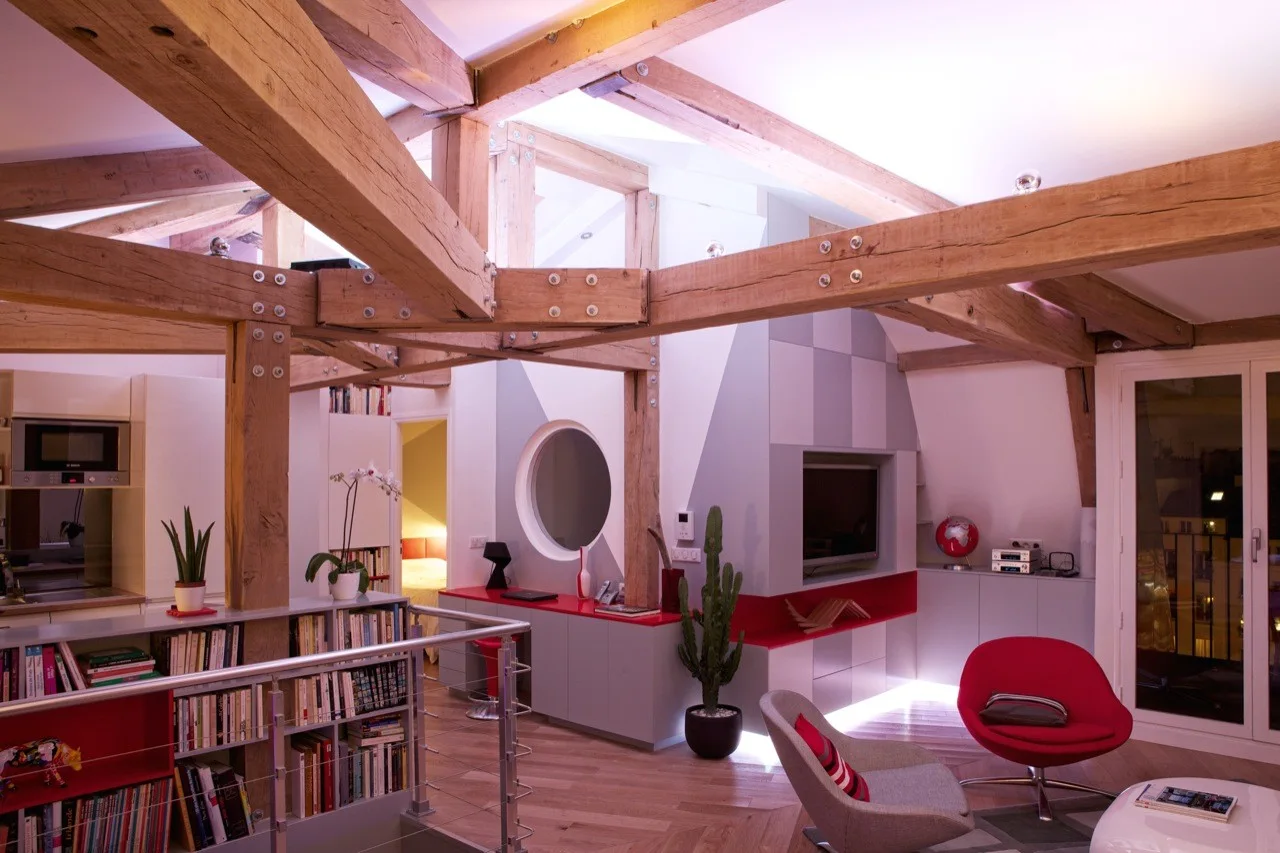 A modern loft living room featuring exposed timber ceiling beams, gray built-in cabinets with red tops, and a red lounge chair.