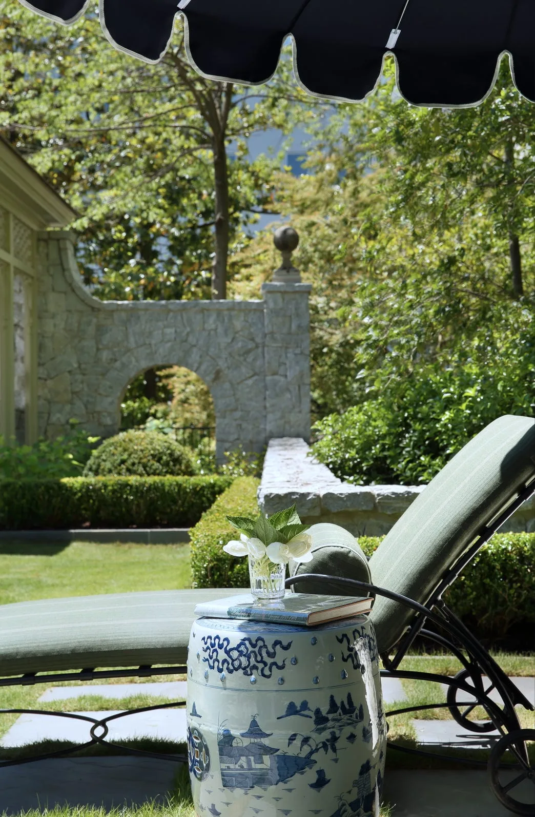 A green upholstered chaise lounge sits next to a blue and white ceramic garden stool on a flagstone patio with a stone archway in the background.