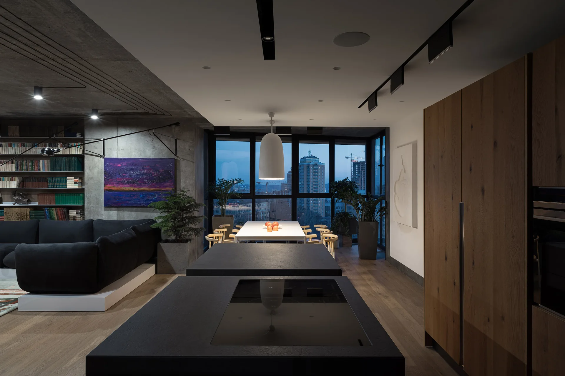 An open-plan apartment features a dark kitchen island, a dining table with a pendant light, and a living area beneath a concrete ceiling.