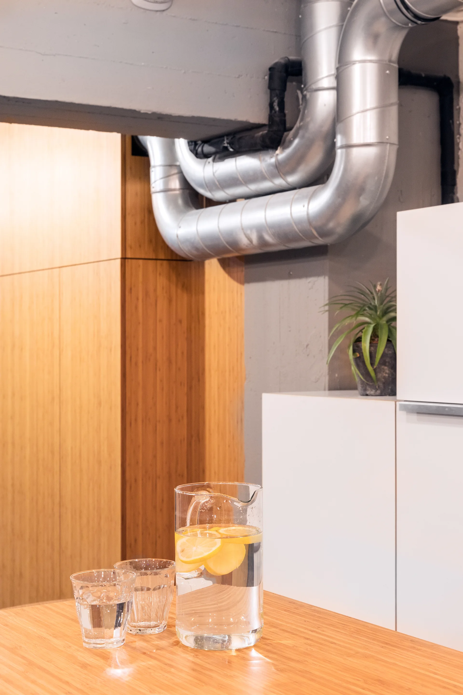 A glass water pitcher with lemon slices sits on a bamboo kitchen table under exposed silver metal HVAC ductwork and white cabinets.