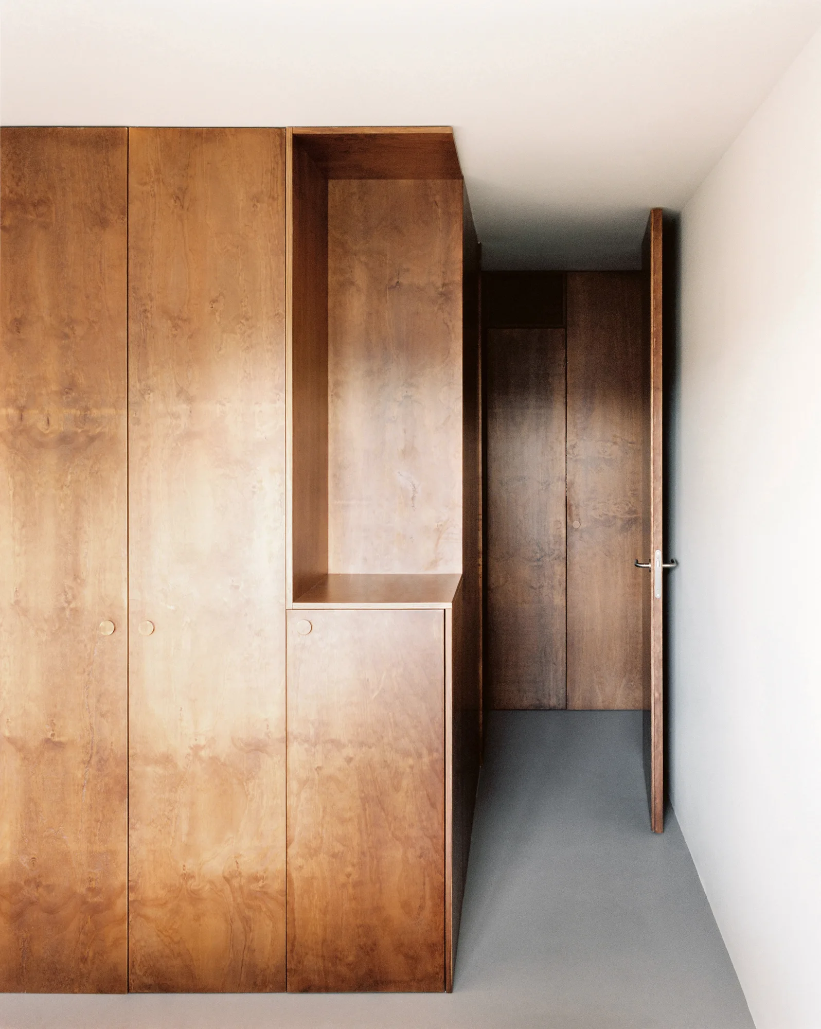 A minimalist hallway featuring floor-to-ceiling wood cabinetry with flush circular pulls, an open niche, off-white walls, and grey concrete floors.