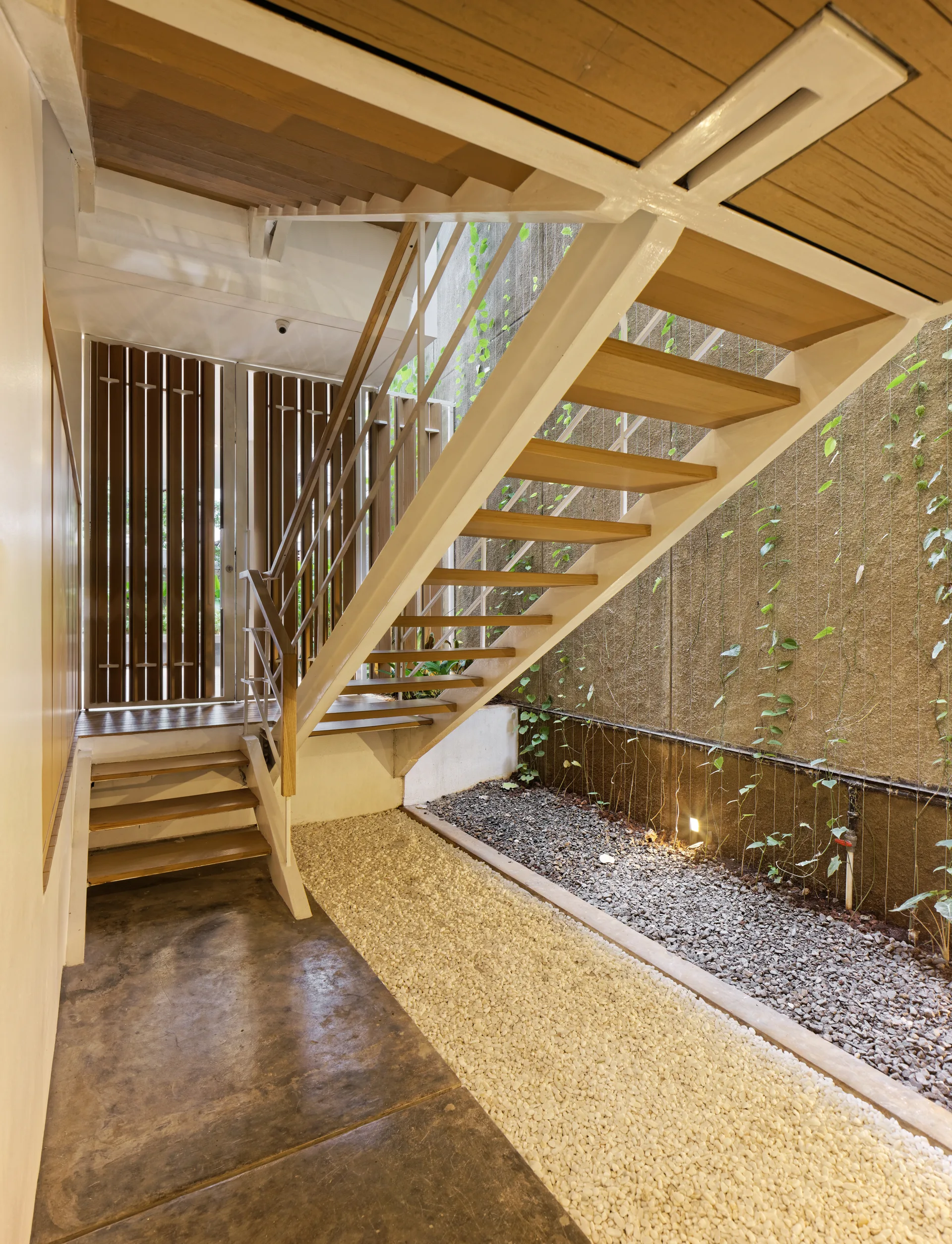 An open riser wooden staircase with white metal supports sits above a gravel garden floor with climbing vines against a textured wall.