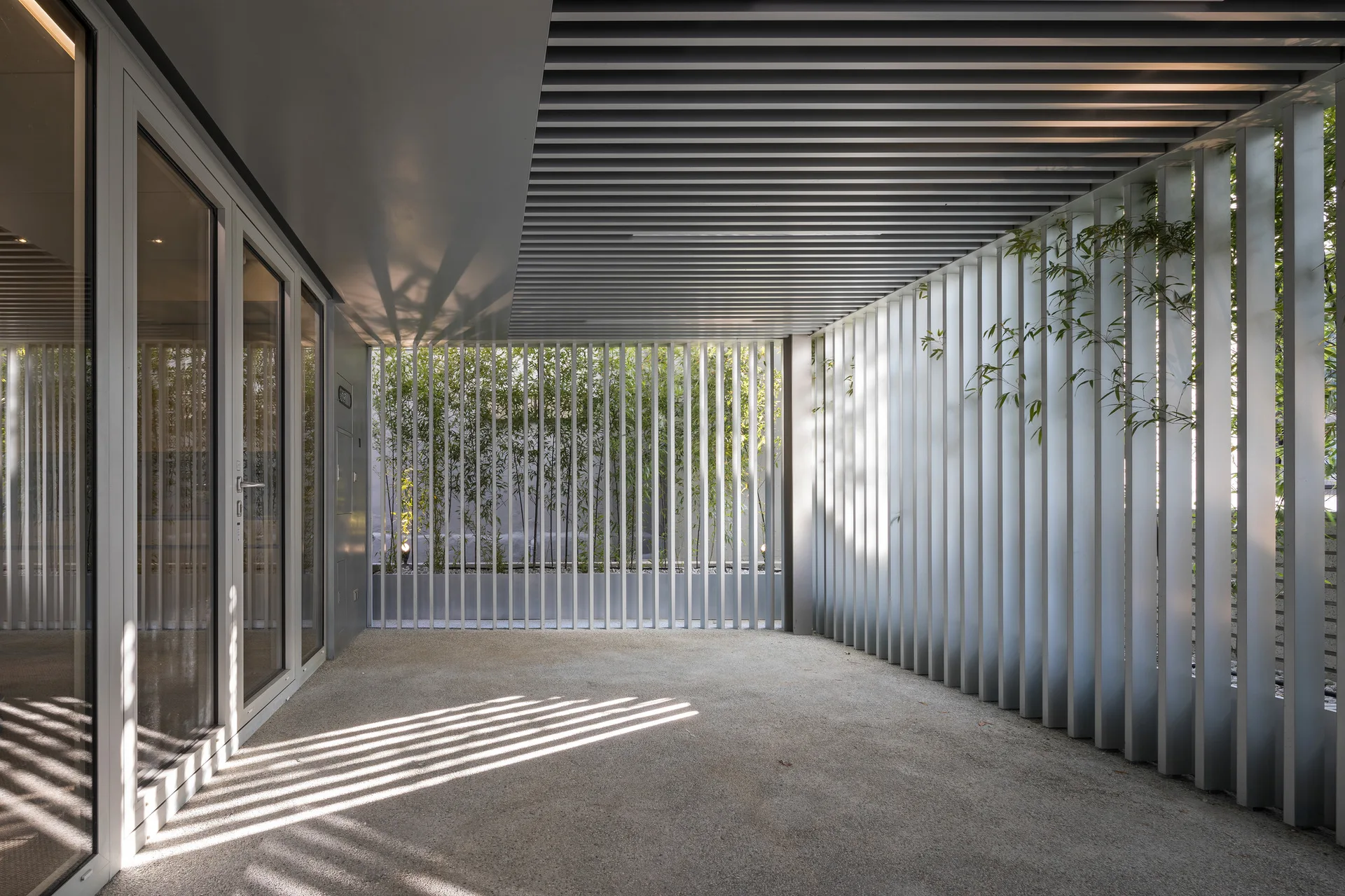 A covered concrete patio features vertical metal slats on the walls, matching ceiling panels, and bamboo plants visible through the screen.