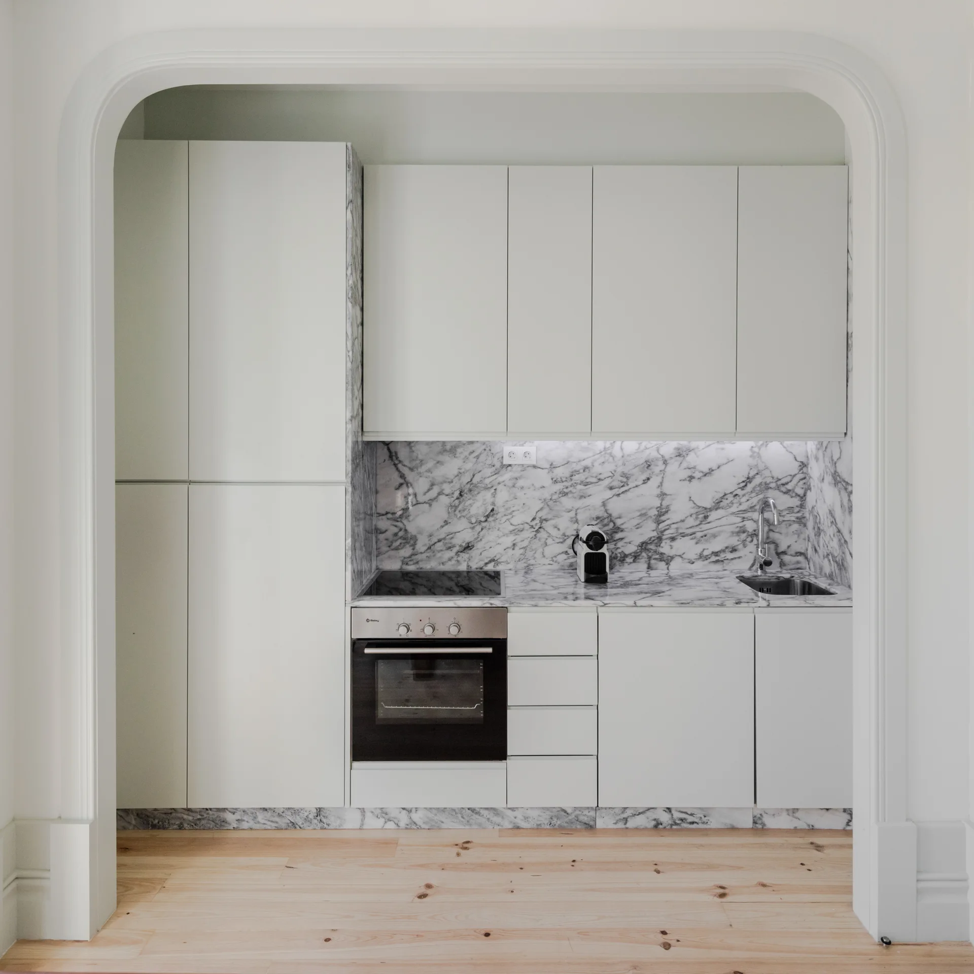 A minimalist kitchen features light grey cabinetry and a heavily veined marble backsplash seen through a wide plaster archway.
