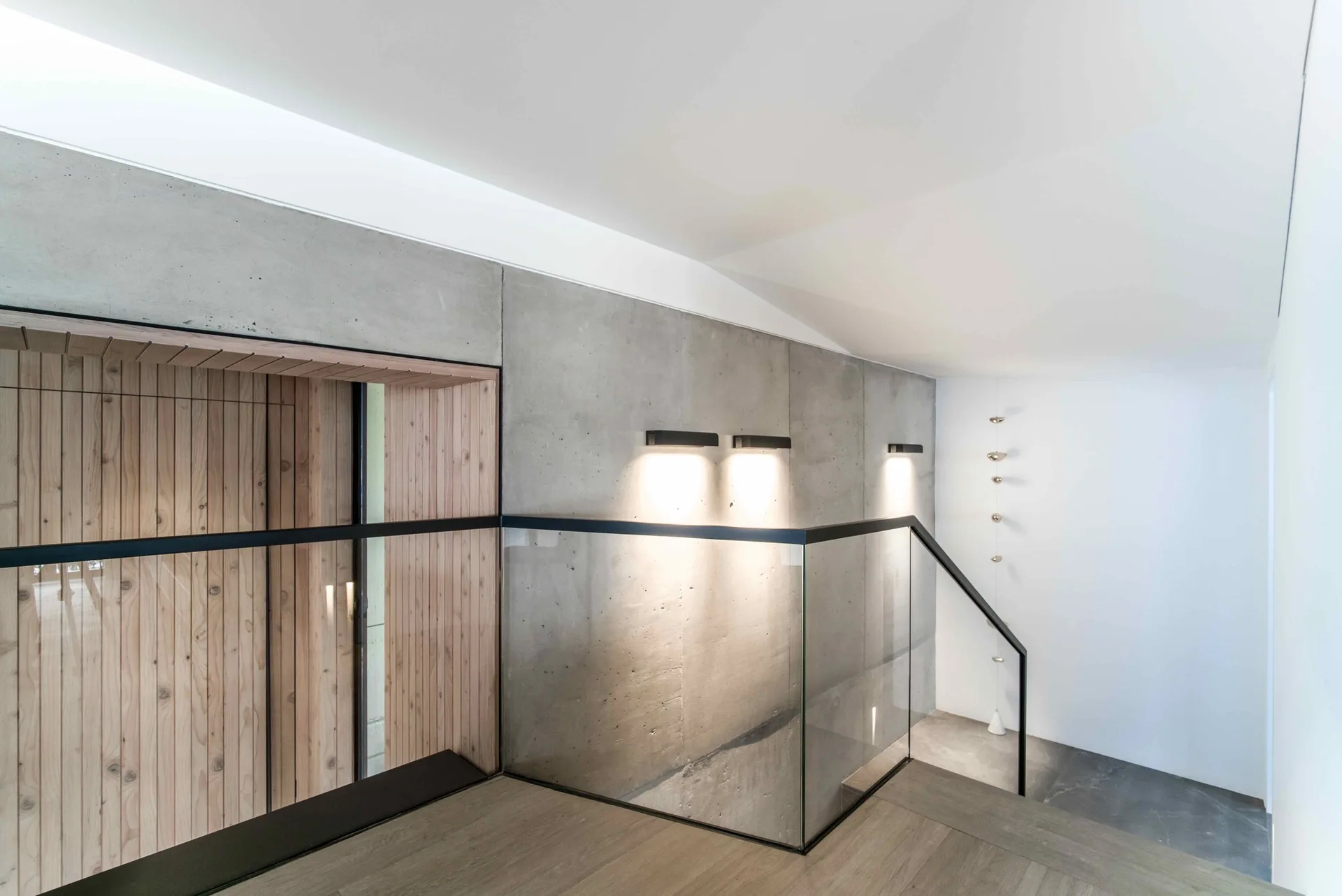 A modern staircase landing features a smooth concrete wall with black sconces, a transparent glass railing, and a recessed wood-paneled niche.