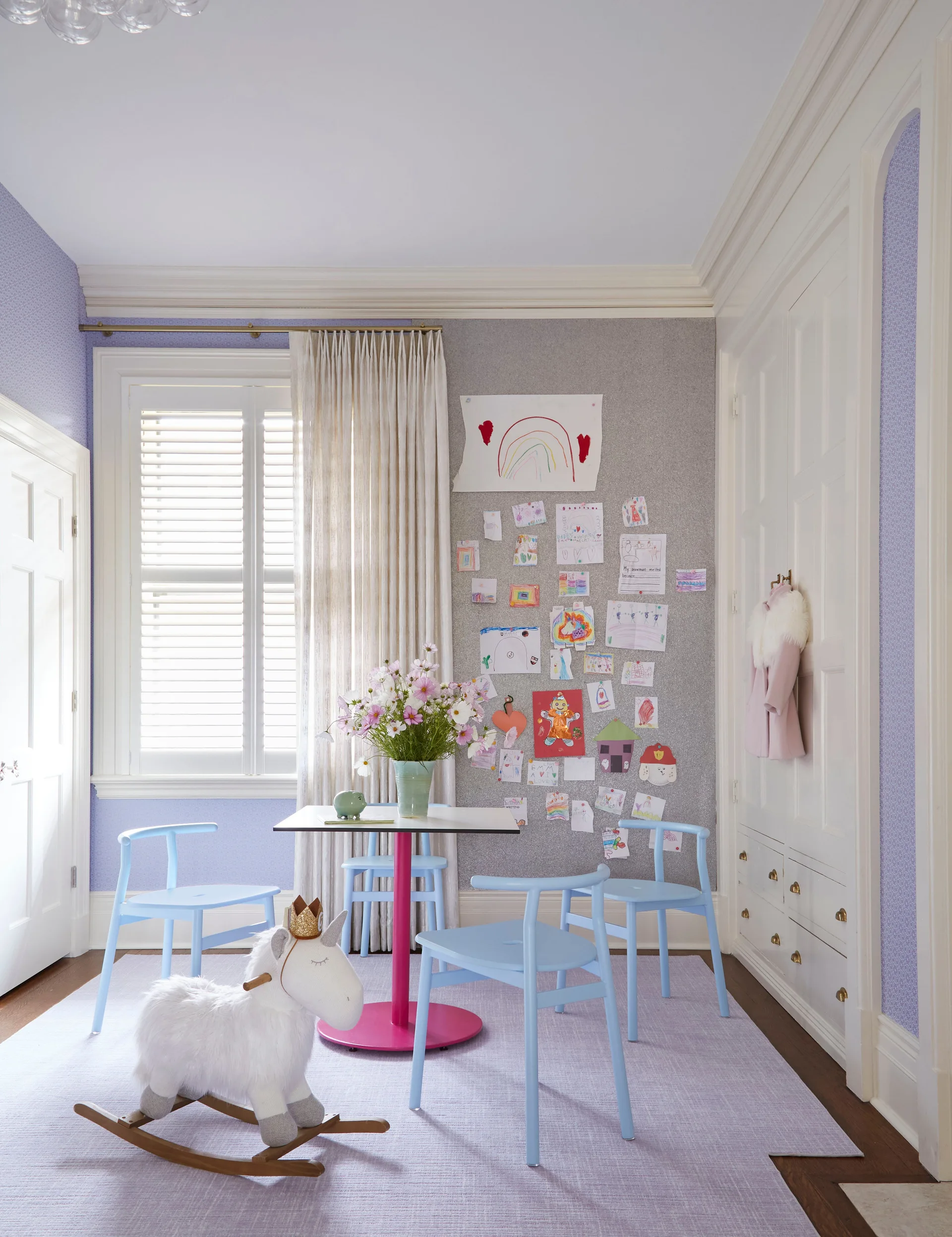 Blue chairs surround a pink-based play table in a lavender kids room with a large pinboard displaying art and built-in white cabinets.