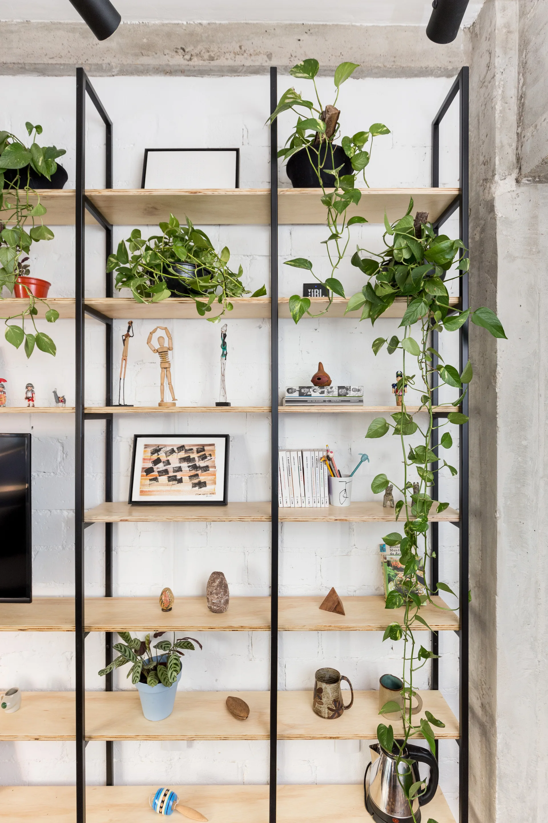 A tall open shelving unit with a black metal frame and light wood shelves stands against a textured white painted brick wall.