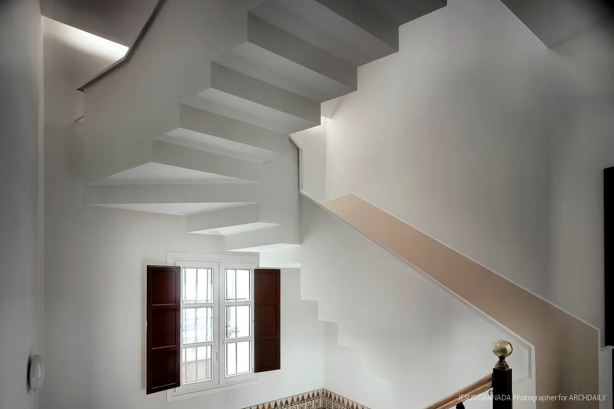 A sculptural white staircase with geometric steps rises next to a small window with open dark wood shutters and a patterned tile baseboard.