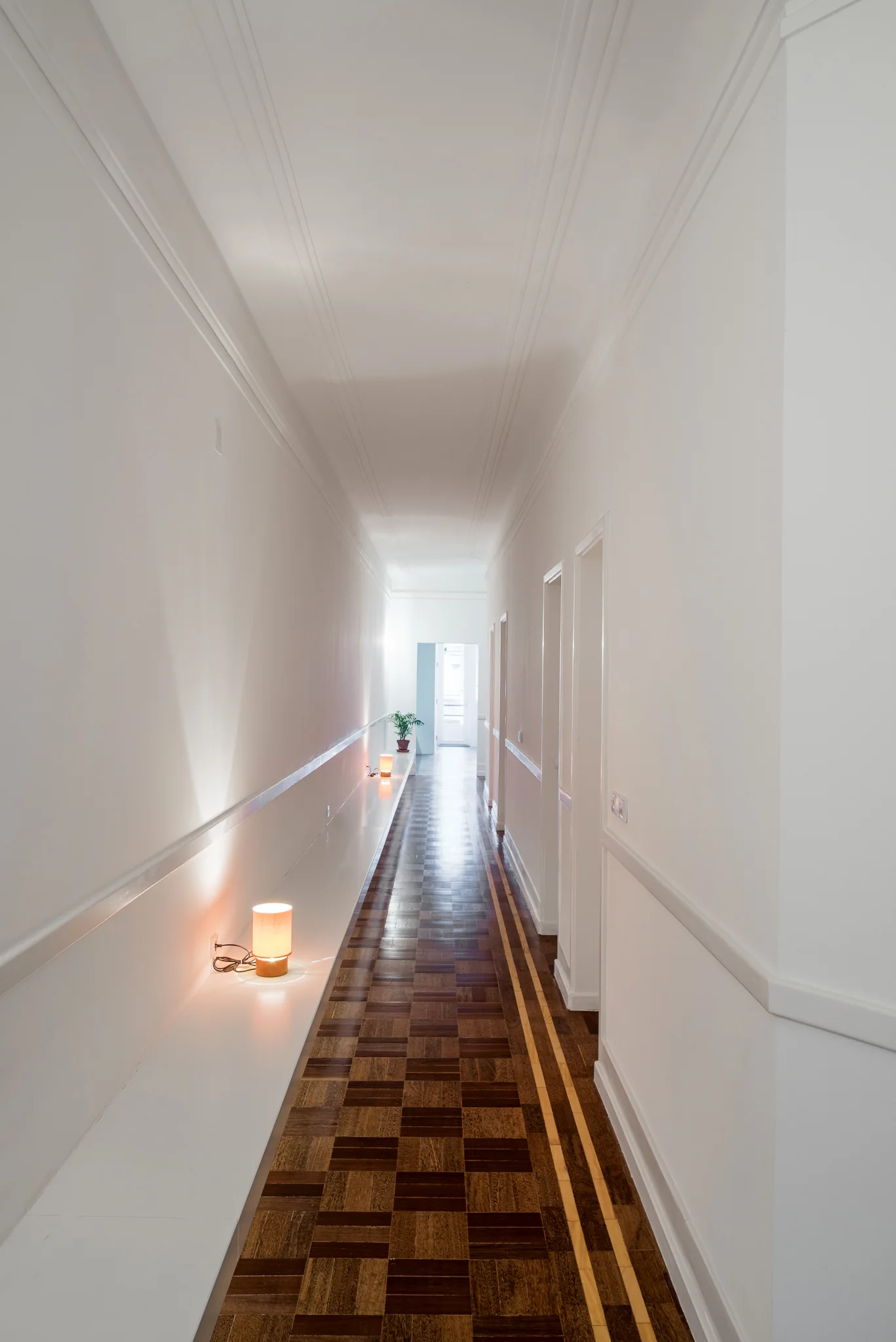 A long white corridor featuring geometric parquet wood flooring, a low built-in shelf with warm cylinder lamps, and several recessed doorways.
