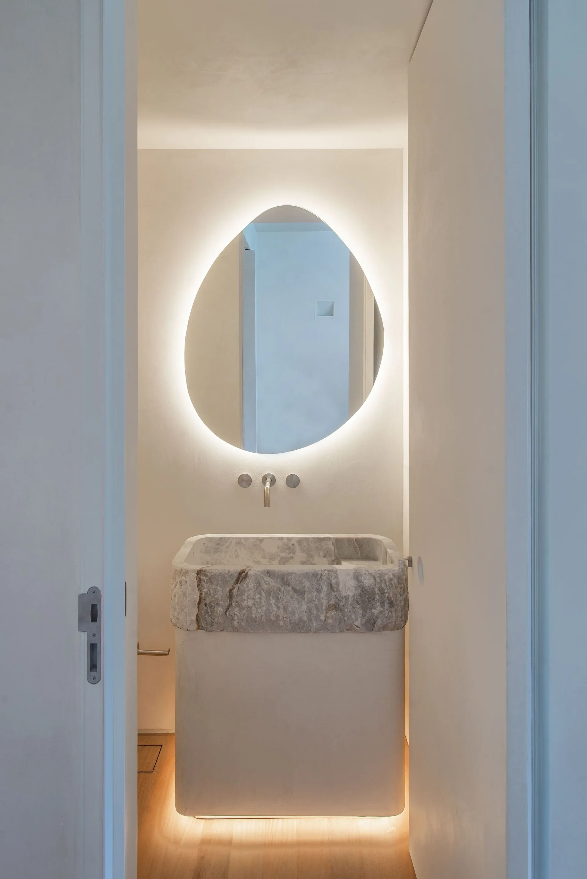 A view through a doorway into a minimalist powder room featuring a rough stone pedestal vanity and a glowing backlit mirror.