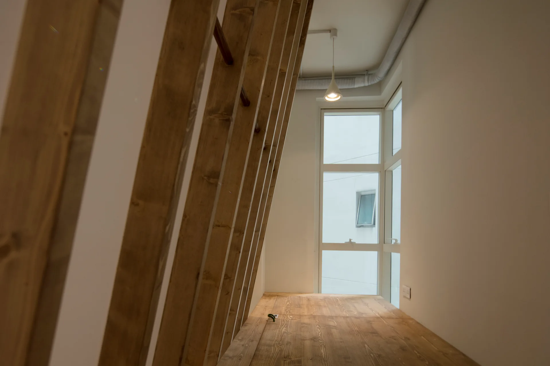 A minimalist room featuring a slanted wood slat partition next to a natural wood floor and a tall window under a single pendant light.