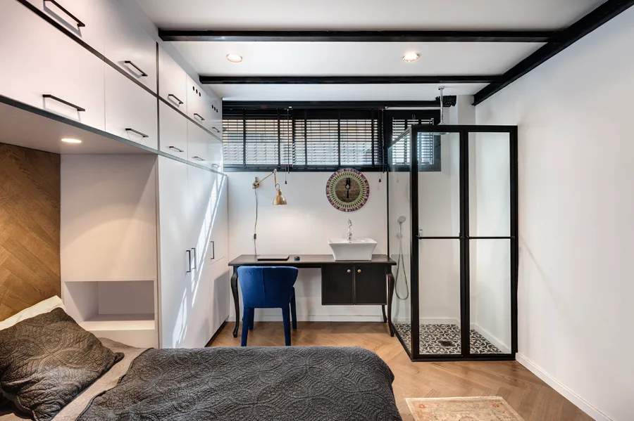 A compact studio bedroom featuring a glass shower with black framing, integrated white wardrobes, and a black desk with an integrated sink.