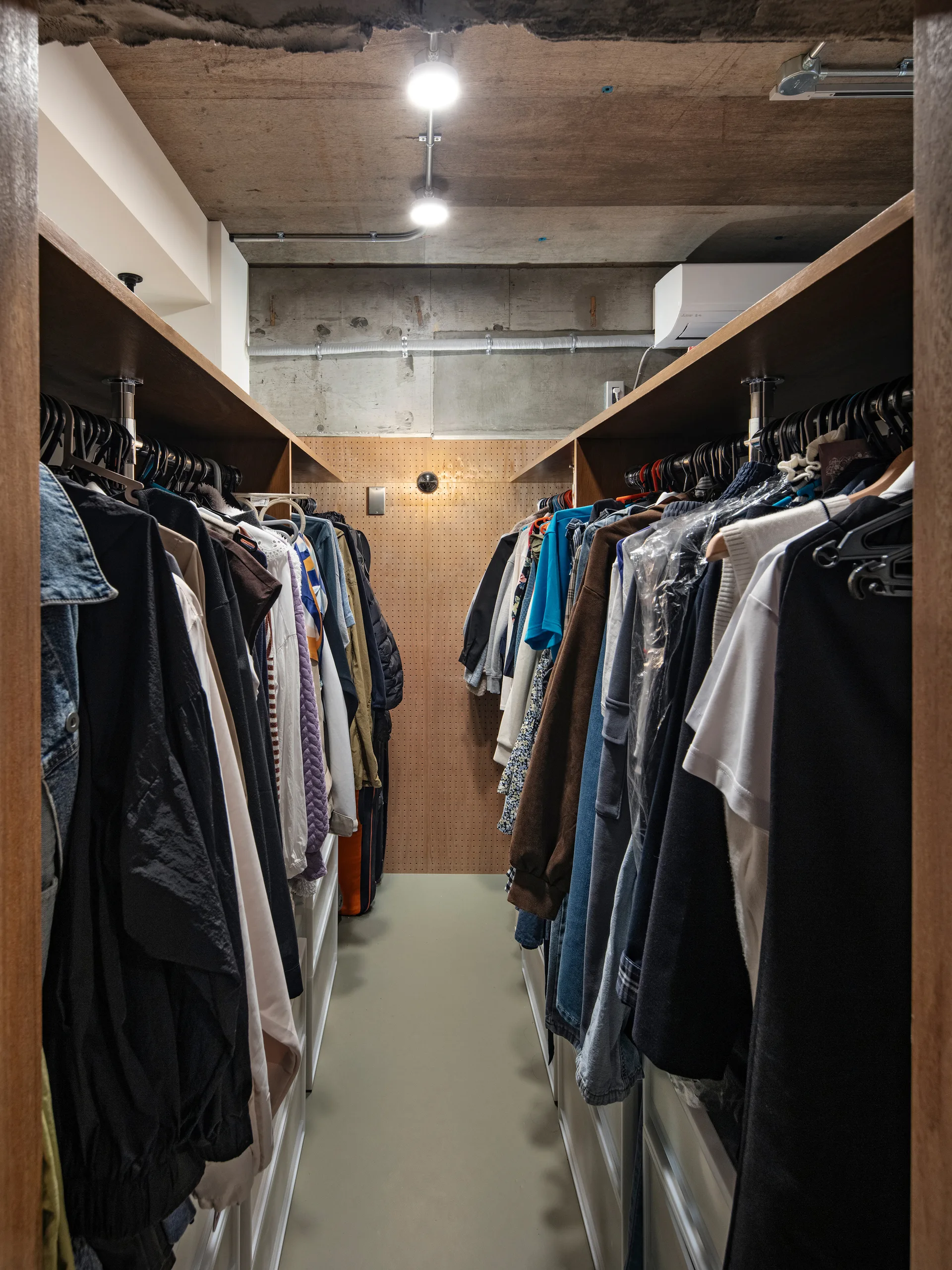 A narrow walk-in closet features clothes hanging on two sides, a wood pegboard back wall, and an exposed concrete ceiling with track lighting.