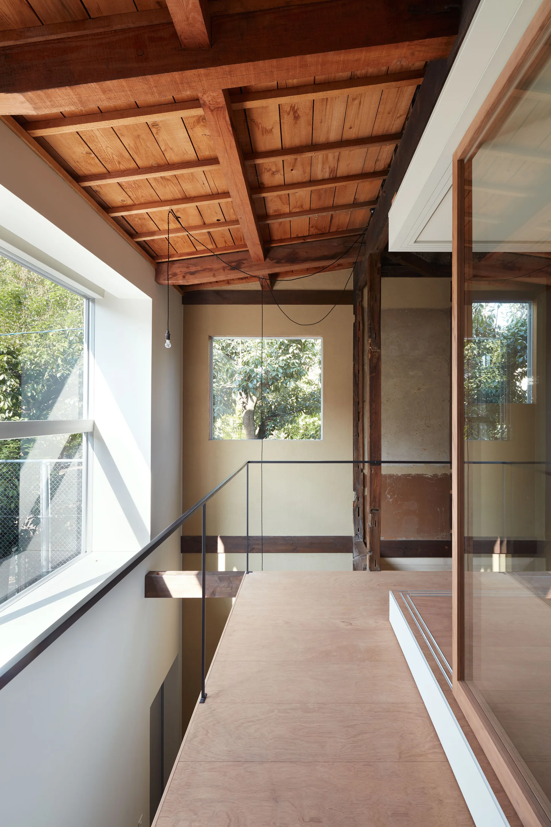 A minimalist wood landing features an exposed timber ceiling, a thin black metal railing, and large windows looking out to green trees.