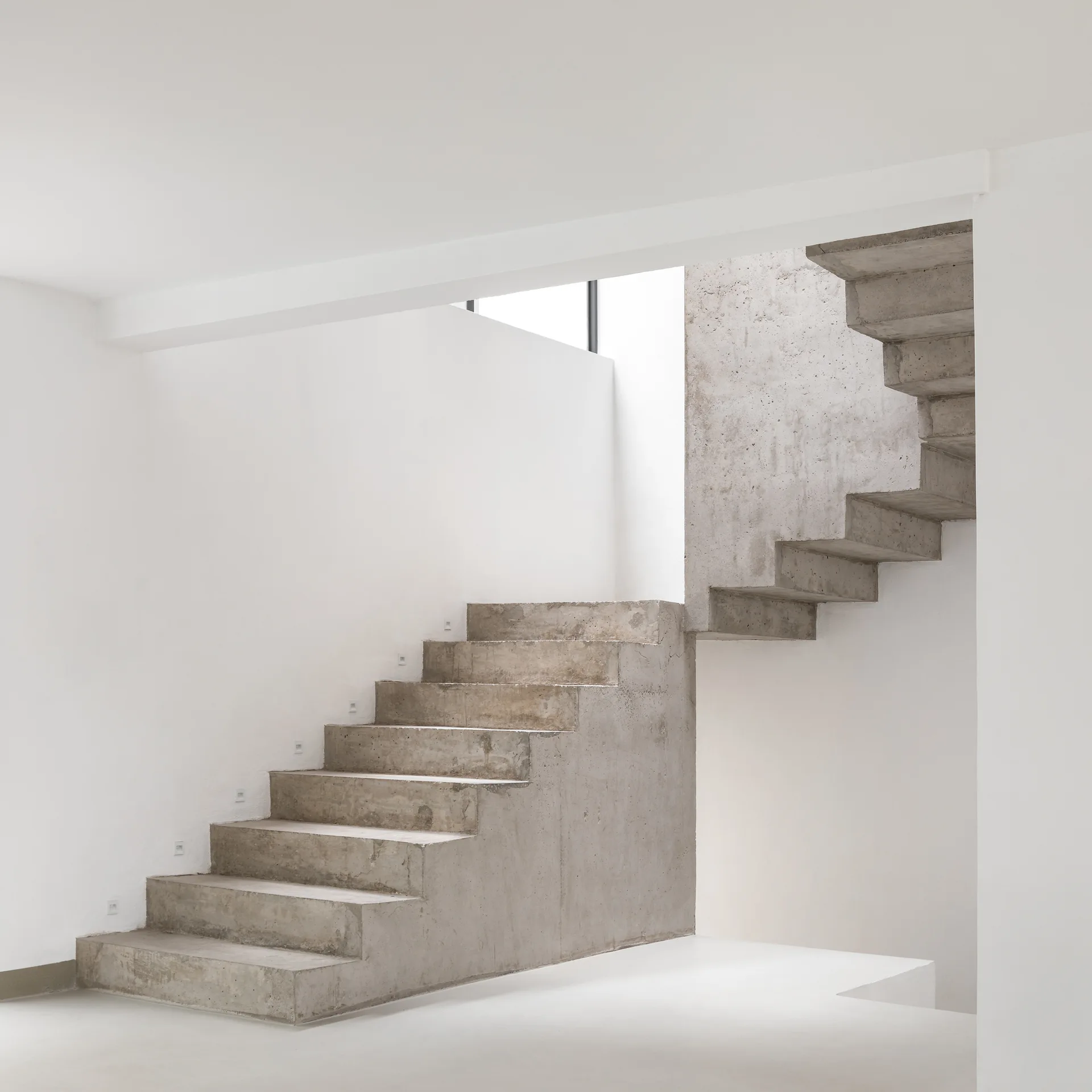 A minimalist concrete staircase rises against clean white walls, featuring small recessed step lights and raw grey concrete textures.