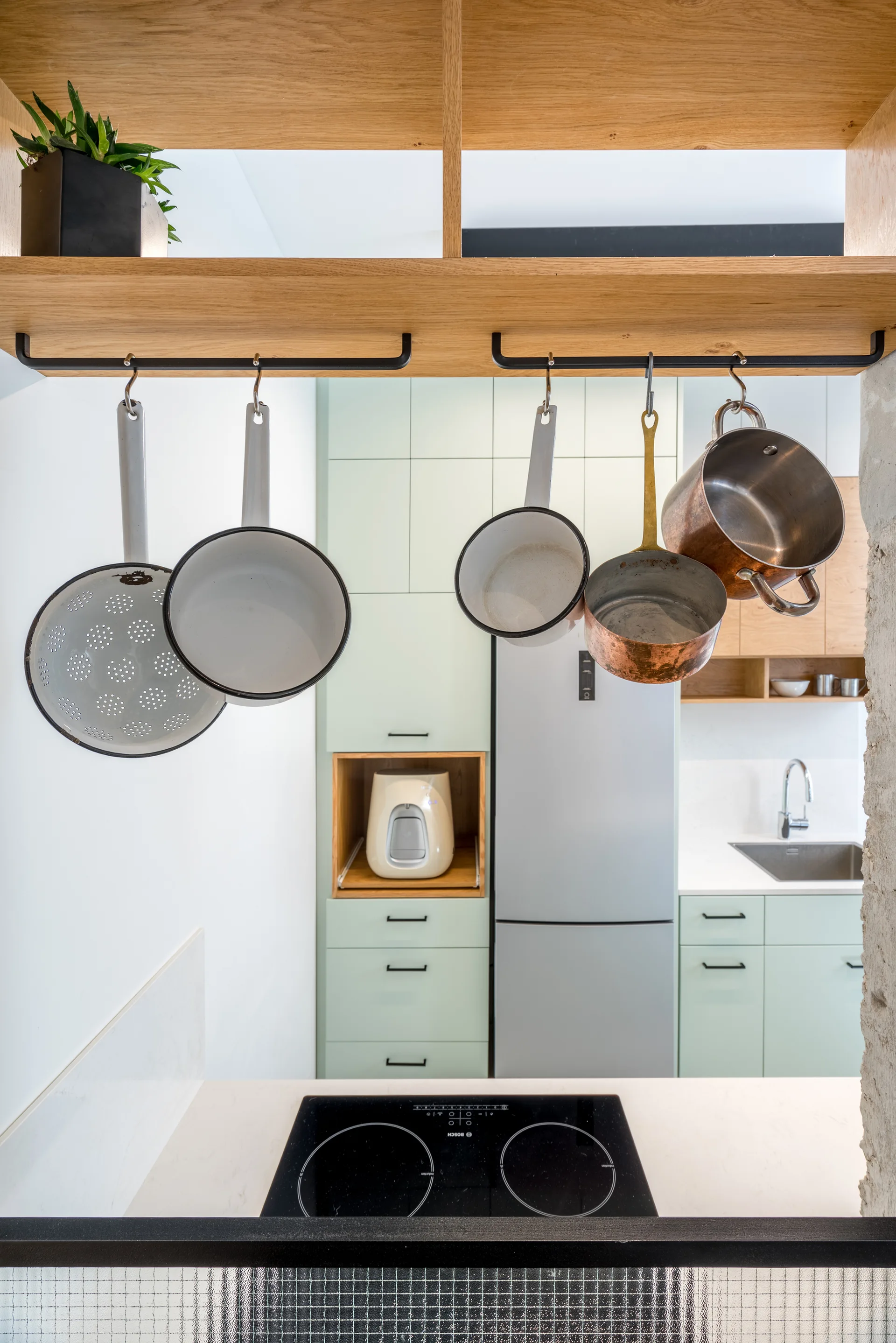 Pots and pans hang from a wooden shelf over an induction cooktop in a kitchen with light green cabinets and a stainless steel refrigerator.