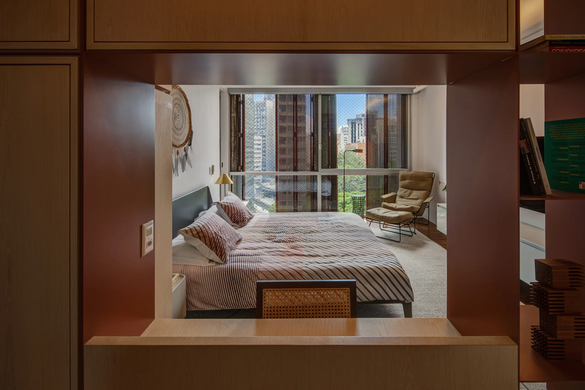 A view through a wood-paneled partition reveals a bedroom with a striped bed, a leather lounge chair, and a large window showing city buildings.
