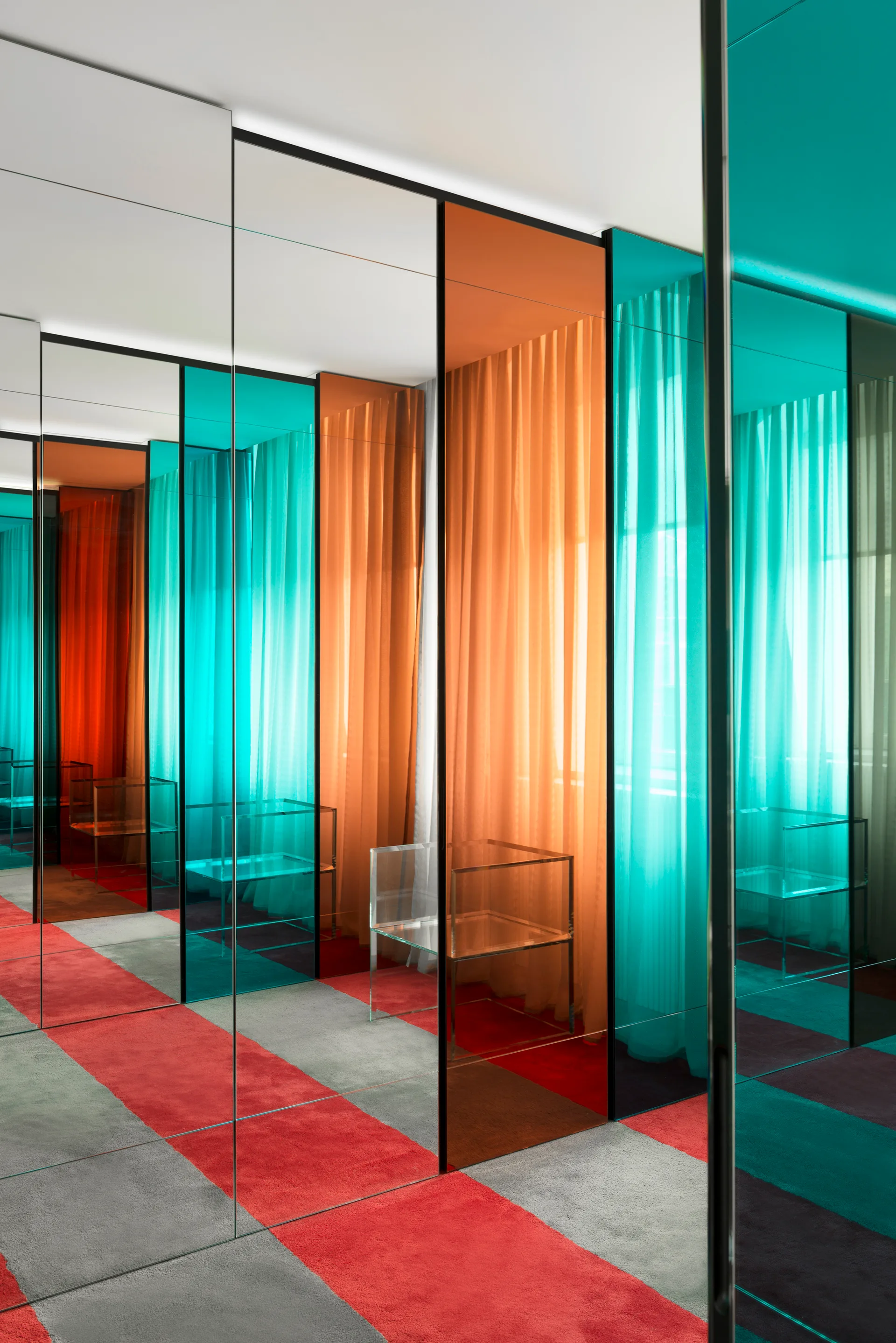 An interior corridor featuring teal and orange colored glass mirror wall panels reflecting transparent acrylic chairs and striped red and grey carpeting.