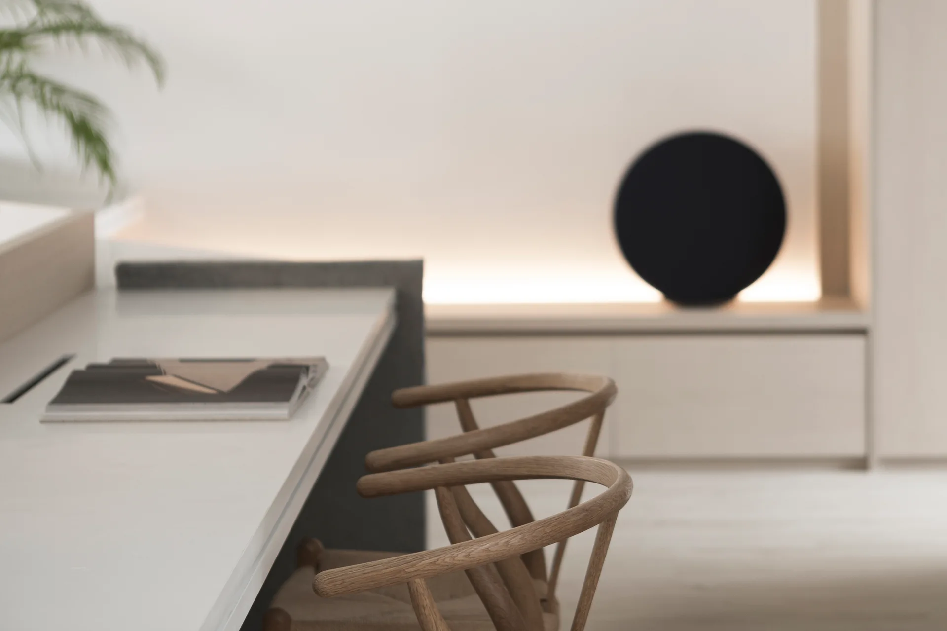 Two wooden wishbone-style chairs sit at a white table in a minimalist beige room with built-in storage and warm cove lighting.