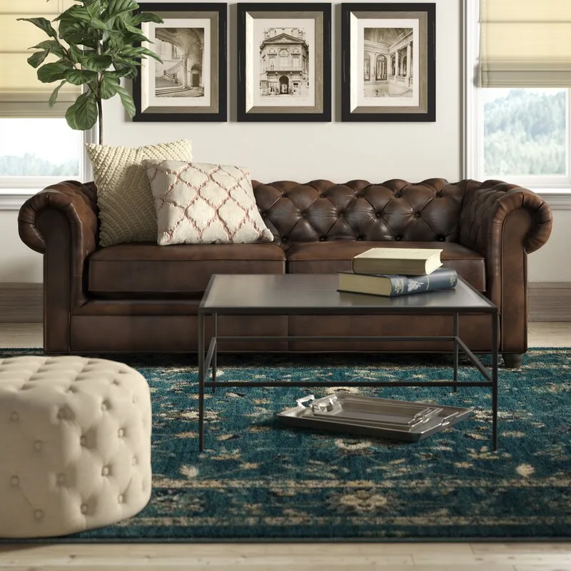 A brown leather Chesterfield sofa sits on a blue patterned rug in front of three framed architectural prints.
