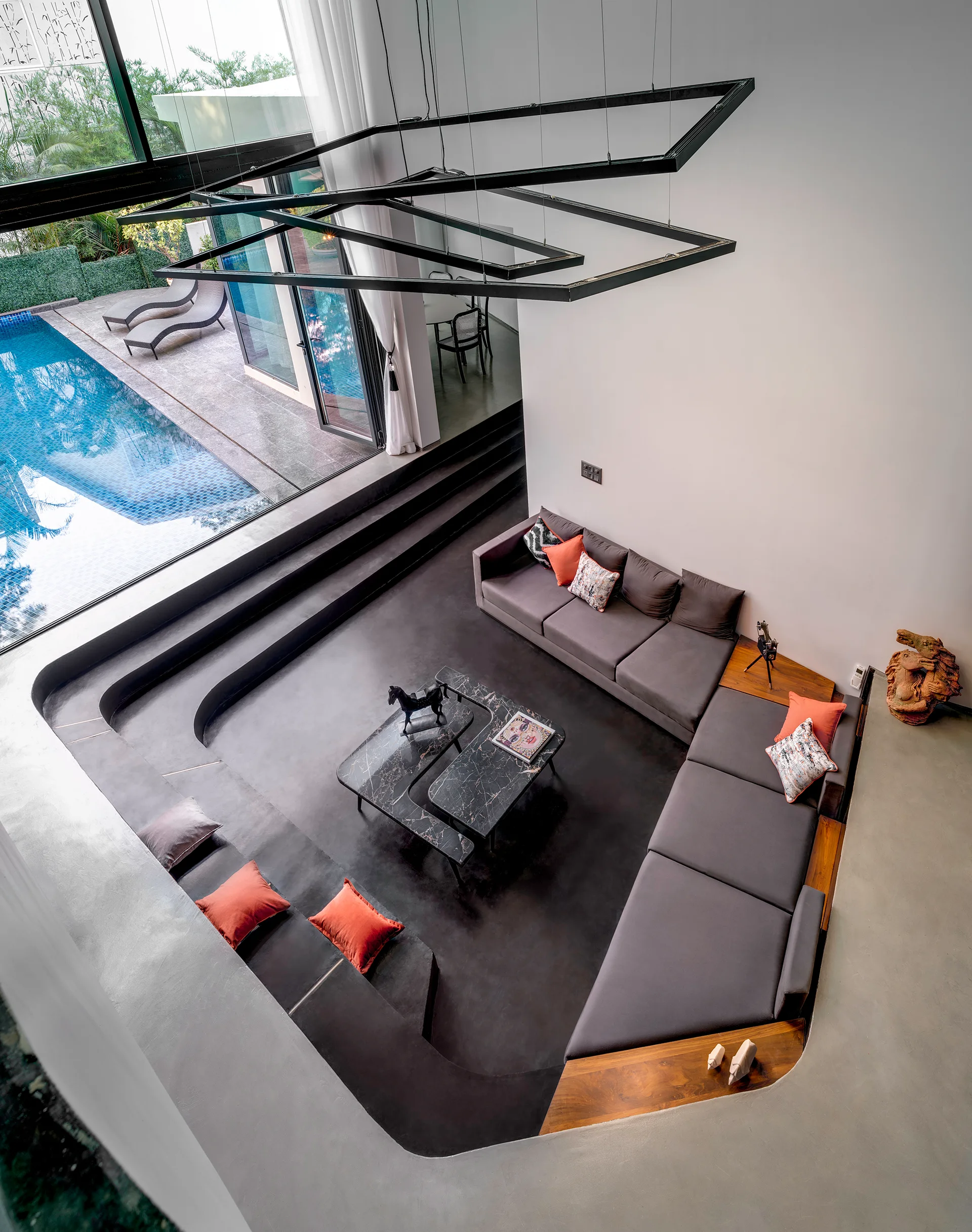 A high-angle view of a modern sunken living room with gray seating, black stone coffee tables, and large windows looking out to a swimming pool.