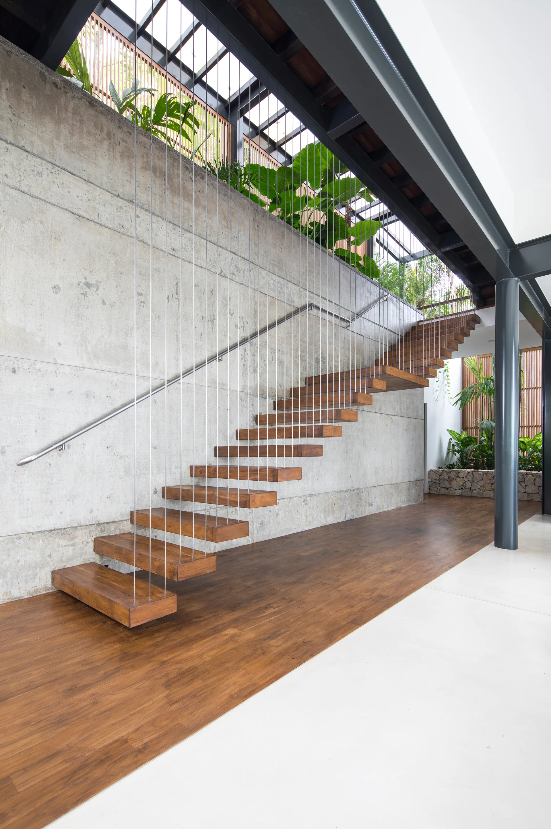 A floating staircase with warm wood treads and vertical steel tension cables is built against a high exposed concrete wall.