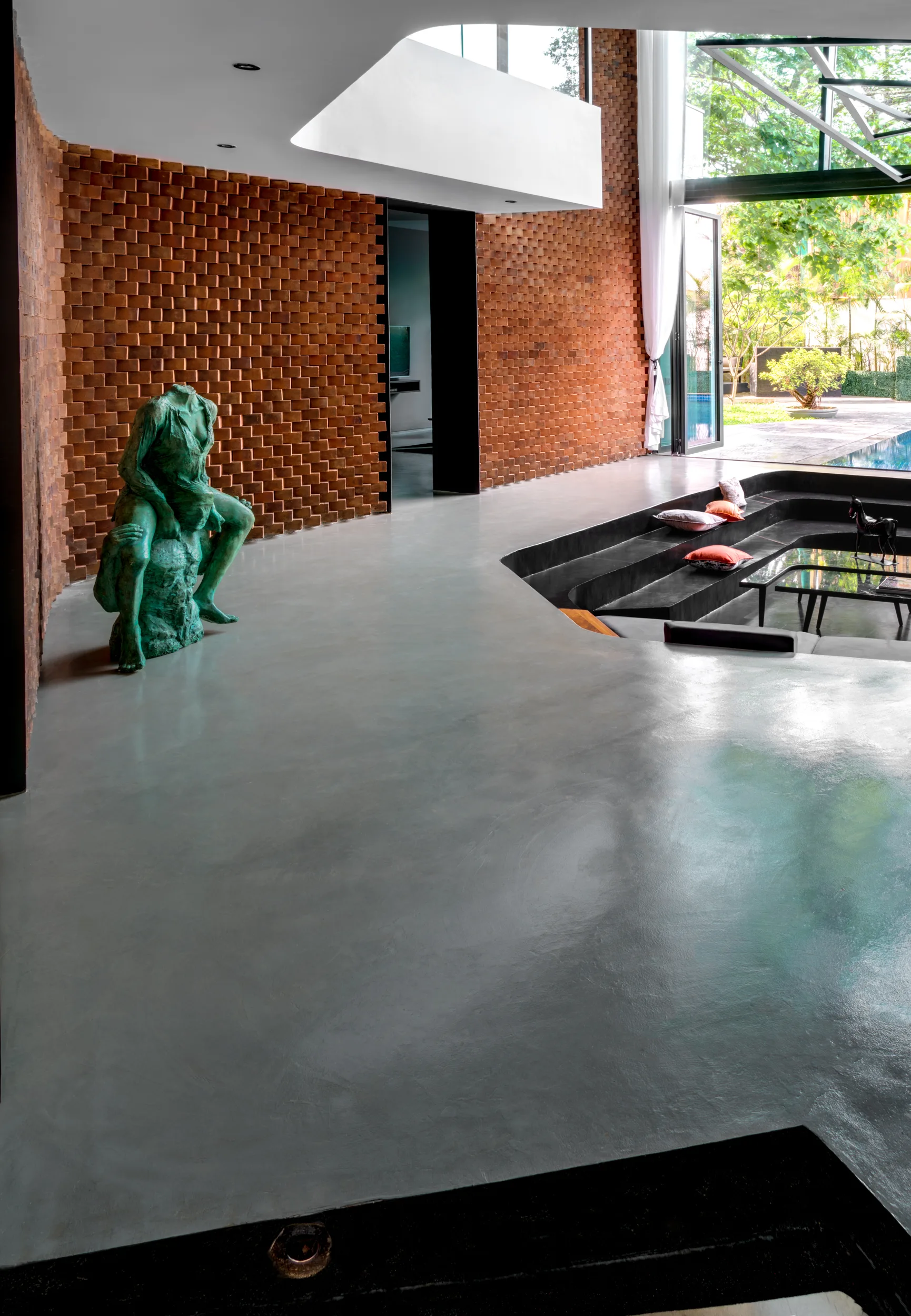 A modern living room features a sunken conversation pit, a textured red brick wall, polished concrete flooring, and a large green sculpture.