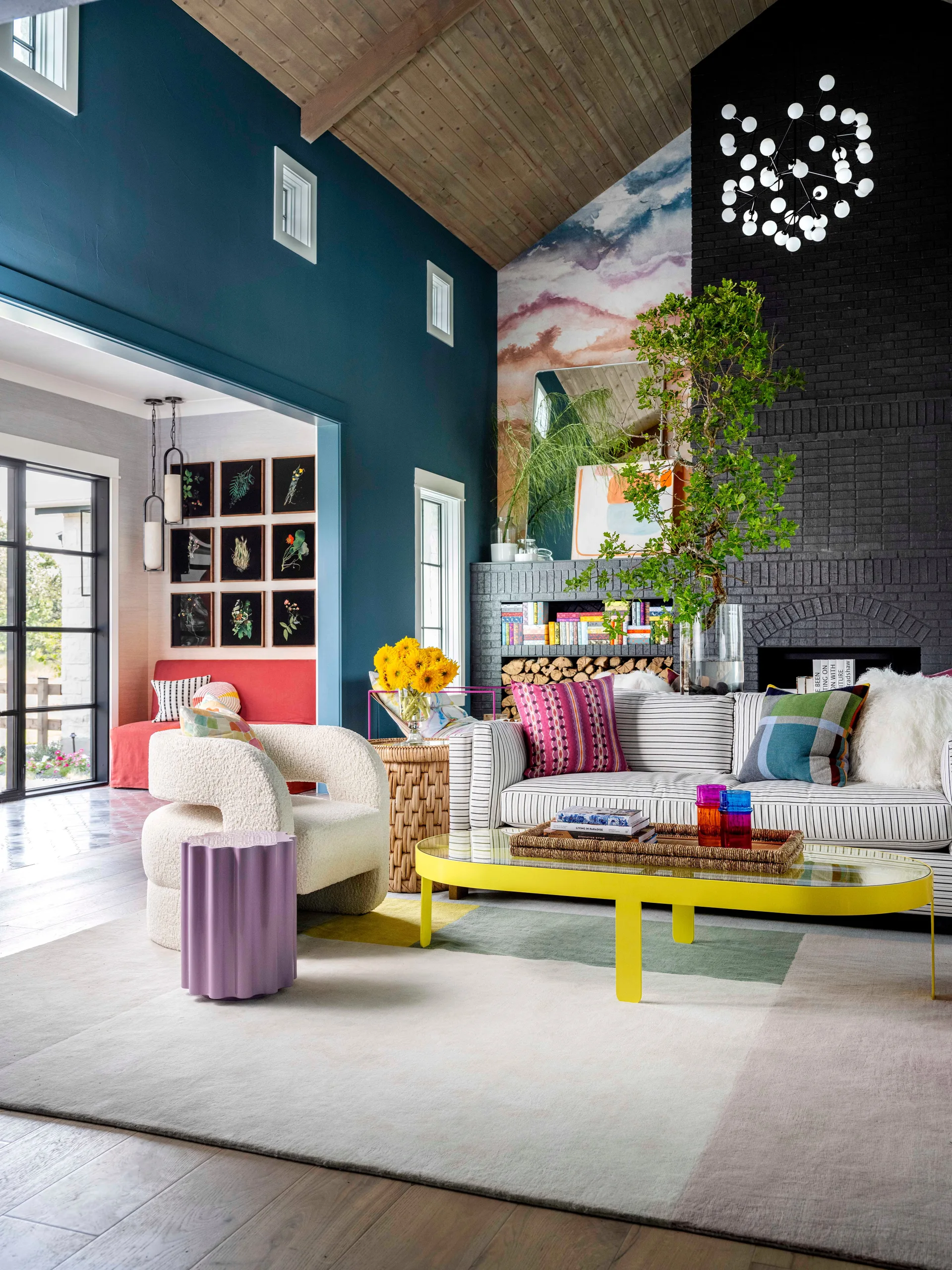 A vibrant living room featuring a striped sofa, a bright yellow coffee table, and a tall black painted brick fireplace.