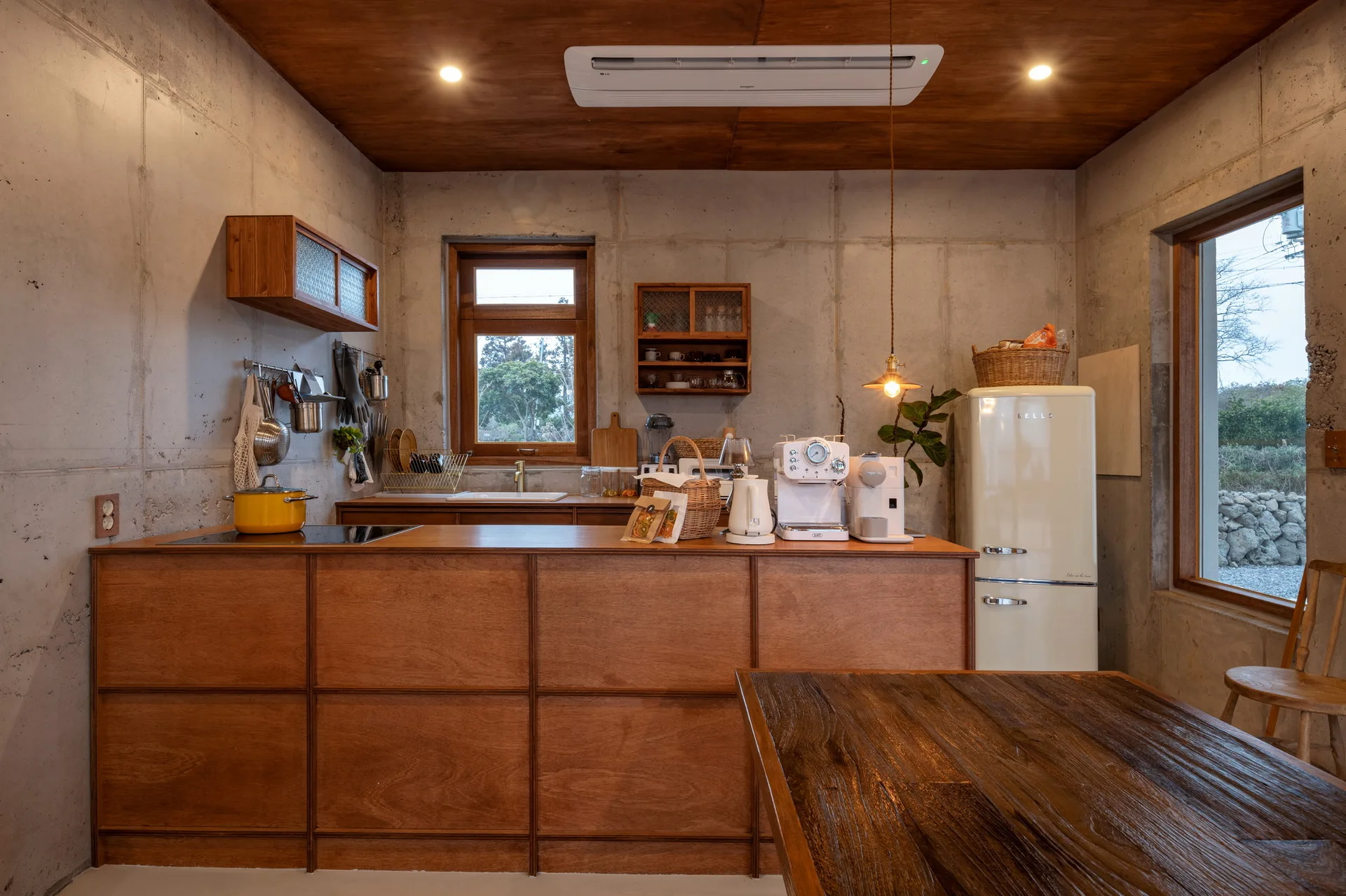 A rustic kitchen features textured grey concrete walls, a warm wood plank ceiling, a matching wooden island, and a cream retro refrigerator.