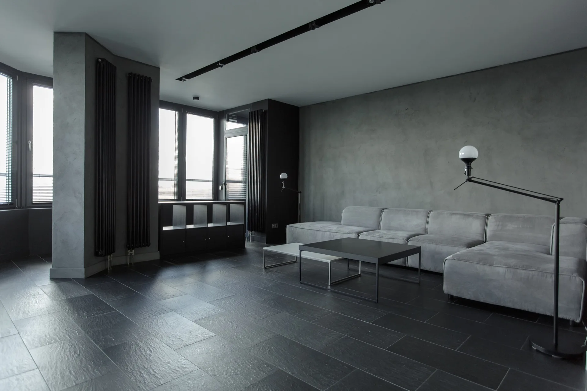 A minimalist grey living room featuring a low-profile sectional sofa, textured plaster walls, black floor tiles, and vertical wall radiators.