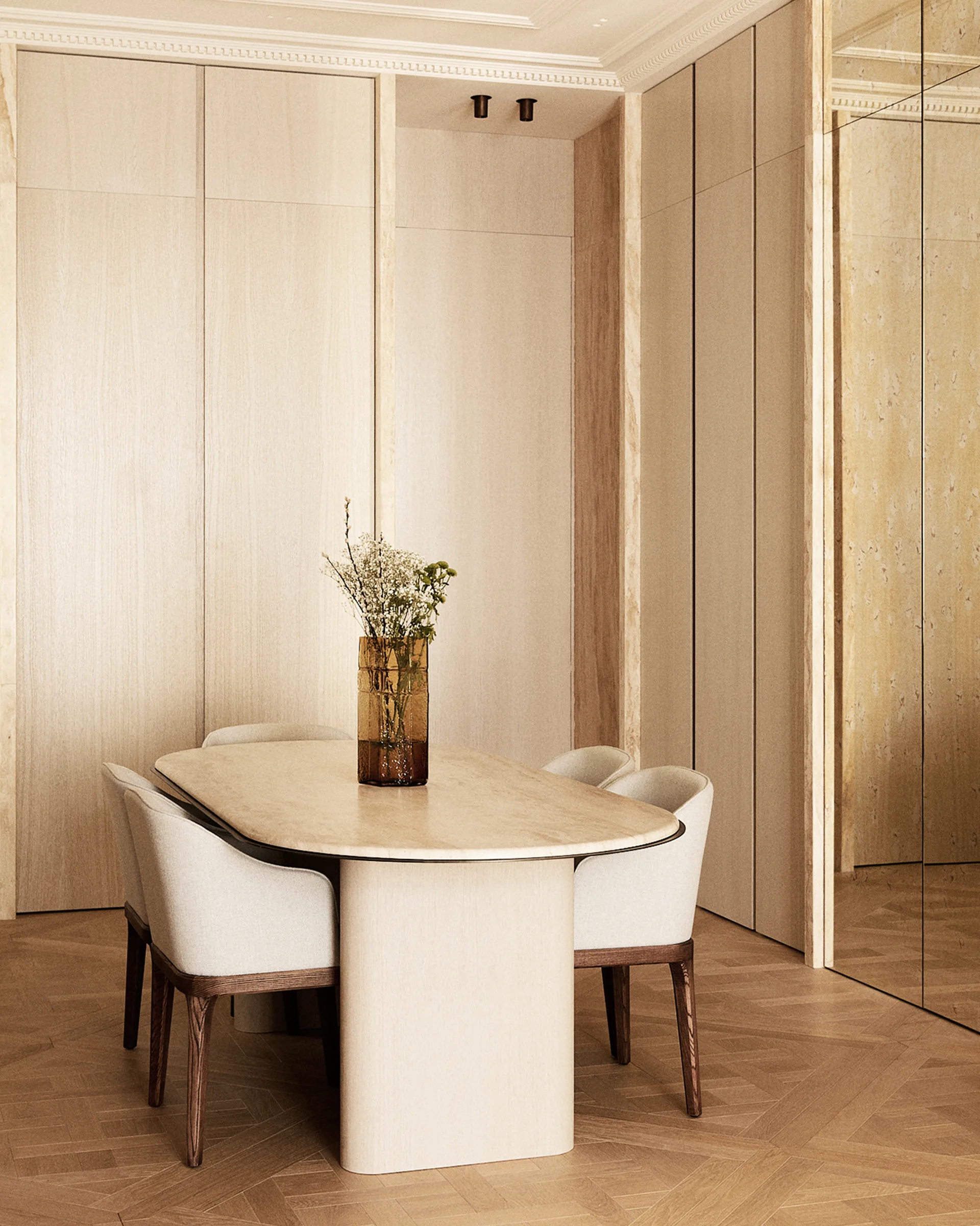 An oval beige stone dining table and four upholstered chairs stand in a room with light wood paneled walls and herringbone flooring.