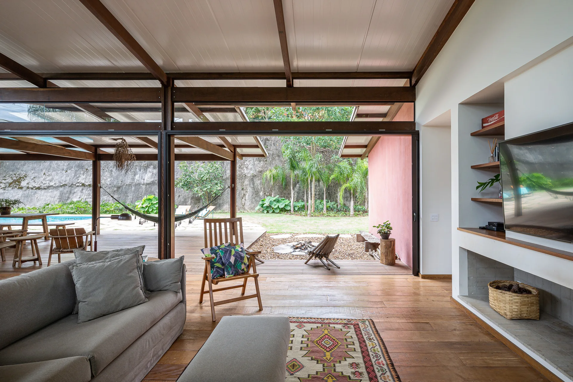 A tropical modern living room with a grey sofa, exposed wood beams, and large open sliding doors leading to an outdoor deck with a hammock.