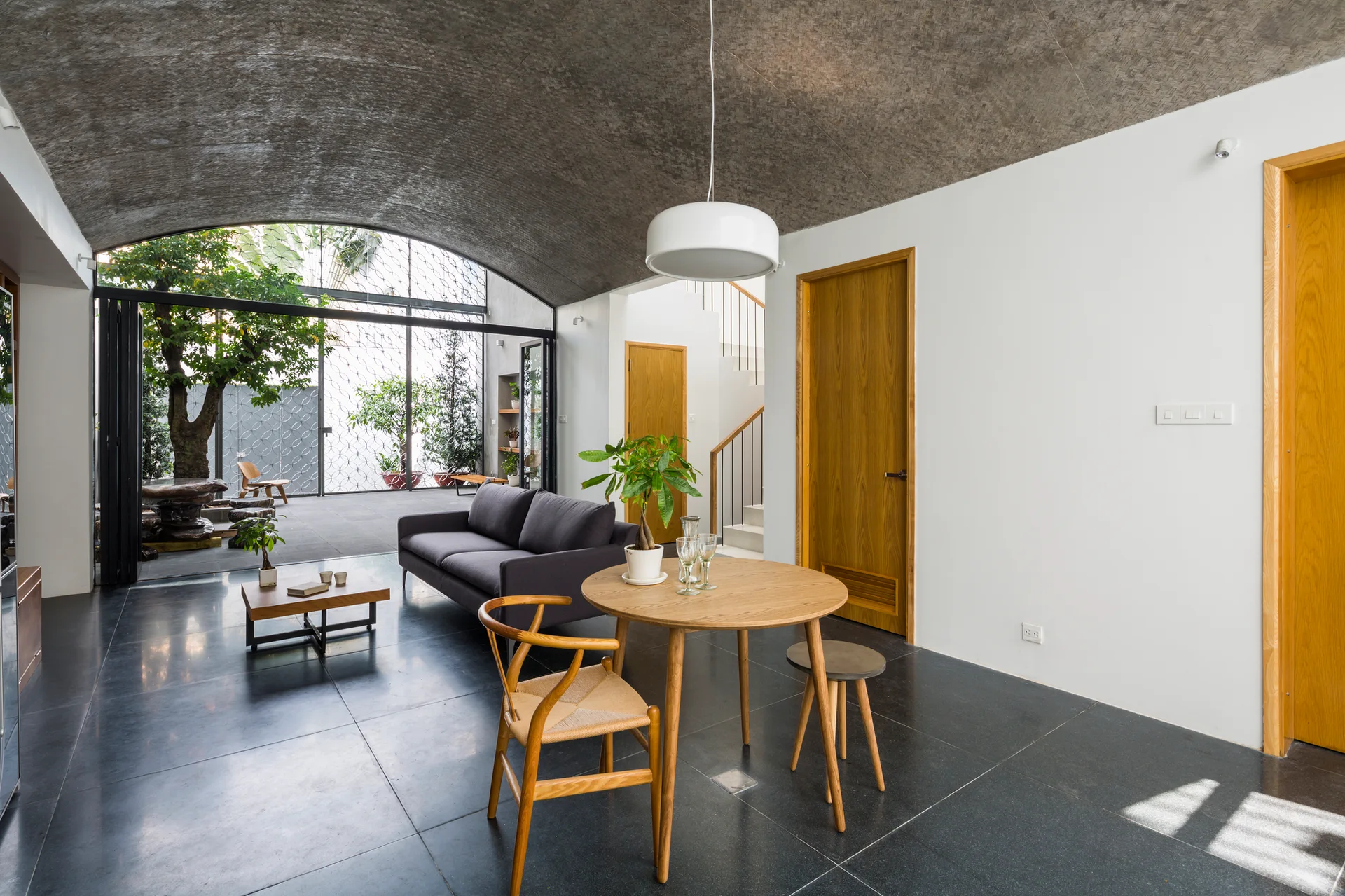 A modern living and dining area features a large textured barrel-vaulted ceiling, dark floor tiles, a round wood table, and a grey sofa.