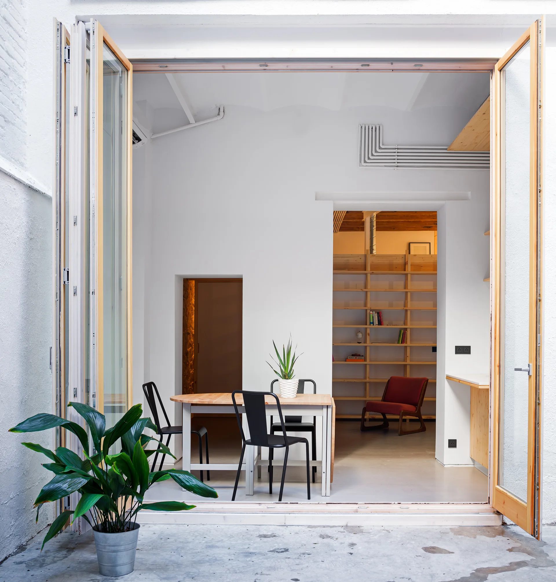 View from a concrete patio through open wood folding doors into a bright dining area with black metal chairs and a light wood table.