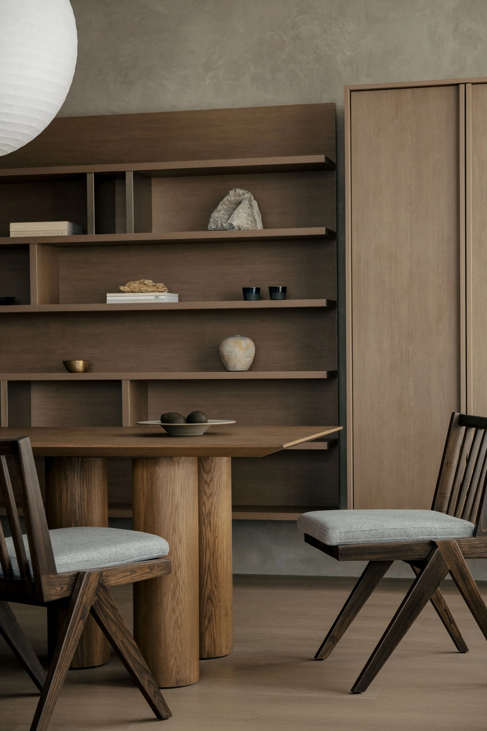 A Japandi dining room features a wooden table with thick cylindrical legs, dark wood chairs with grey cushions, and a large built-in shelving unit.