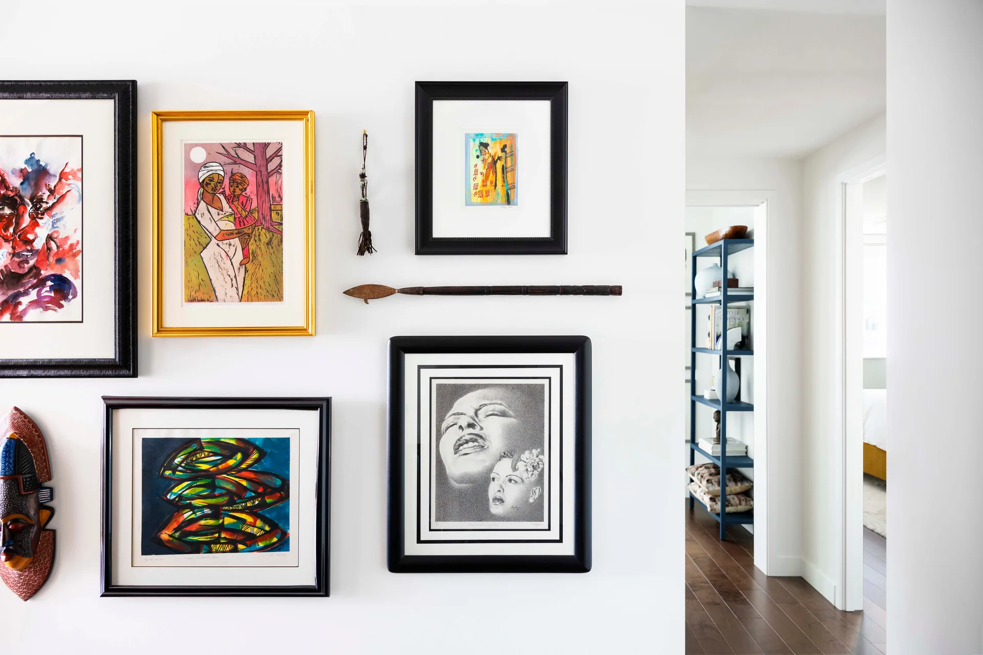 A white wall displays an eclectic gallery arrangement of framed paintings, a carved wood mask, and a small decorative spear.
