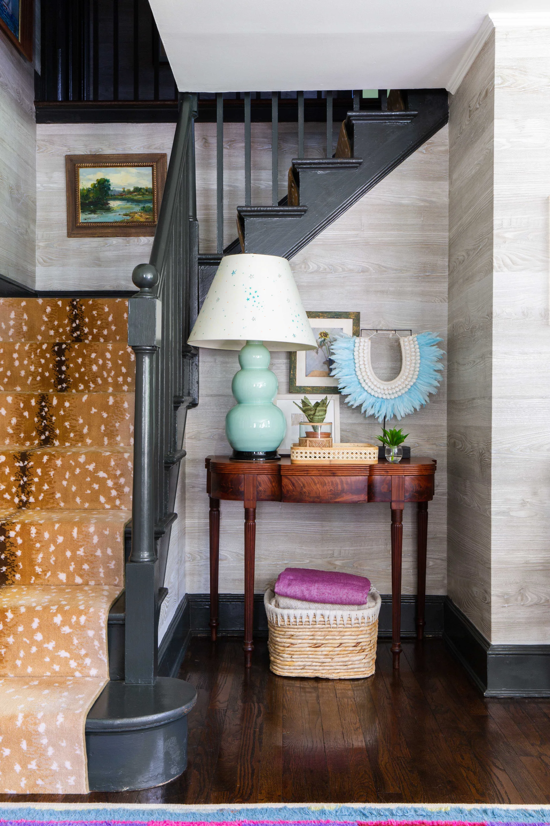 An entryway staircase with a fawn print runner sits next to a dark wood console table displaying an aqua table lamp and decorative items.