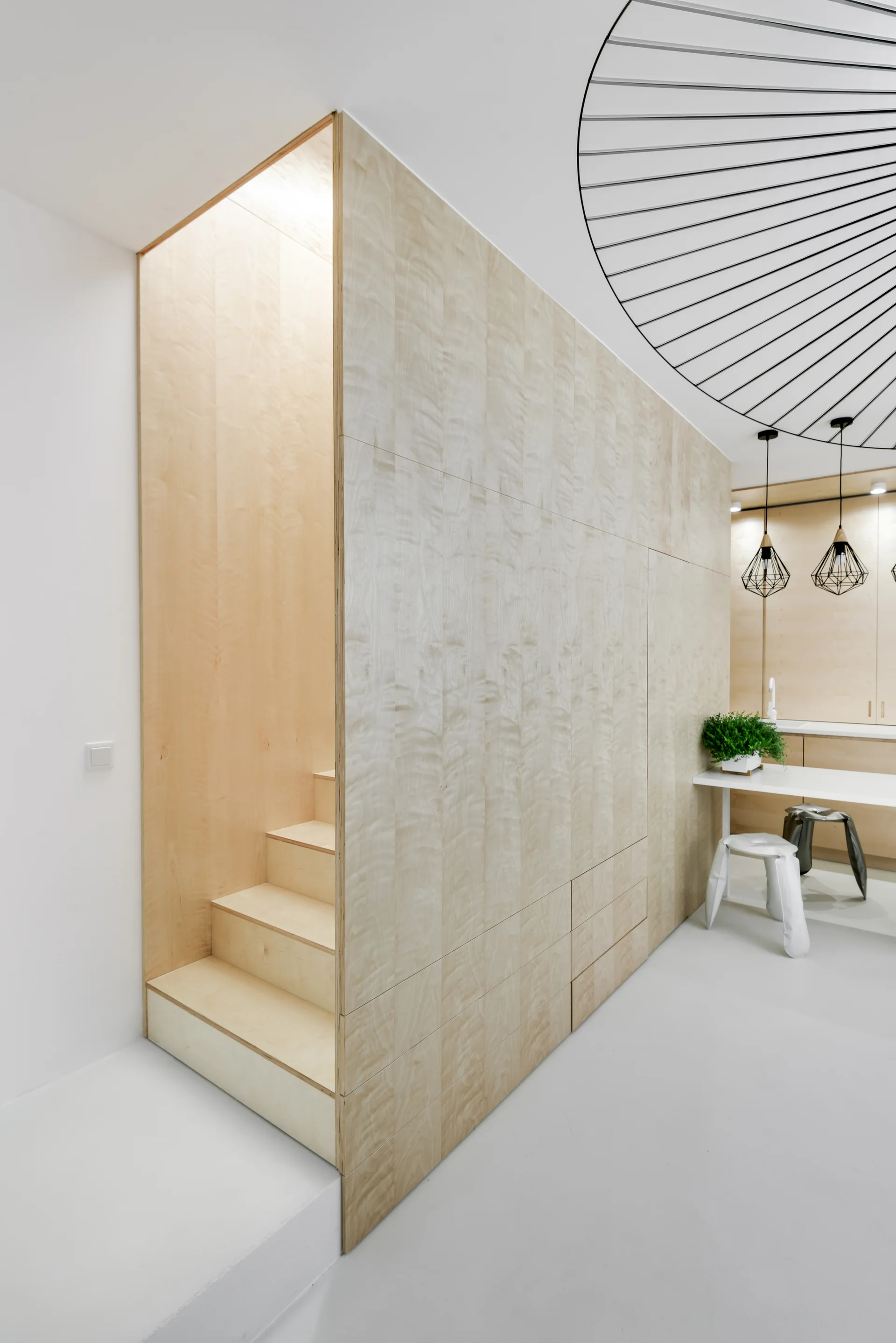 A minimalist white room features a light wood staircase integrated into a seamless wall of built-in cabinetry next to a dining area.