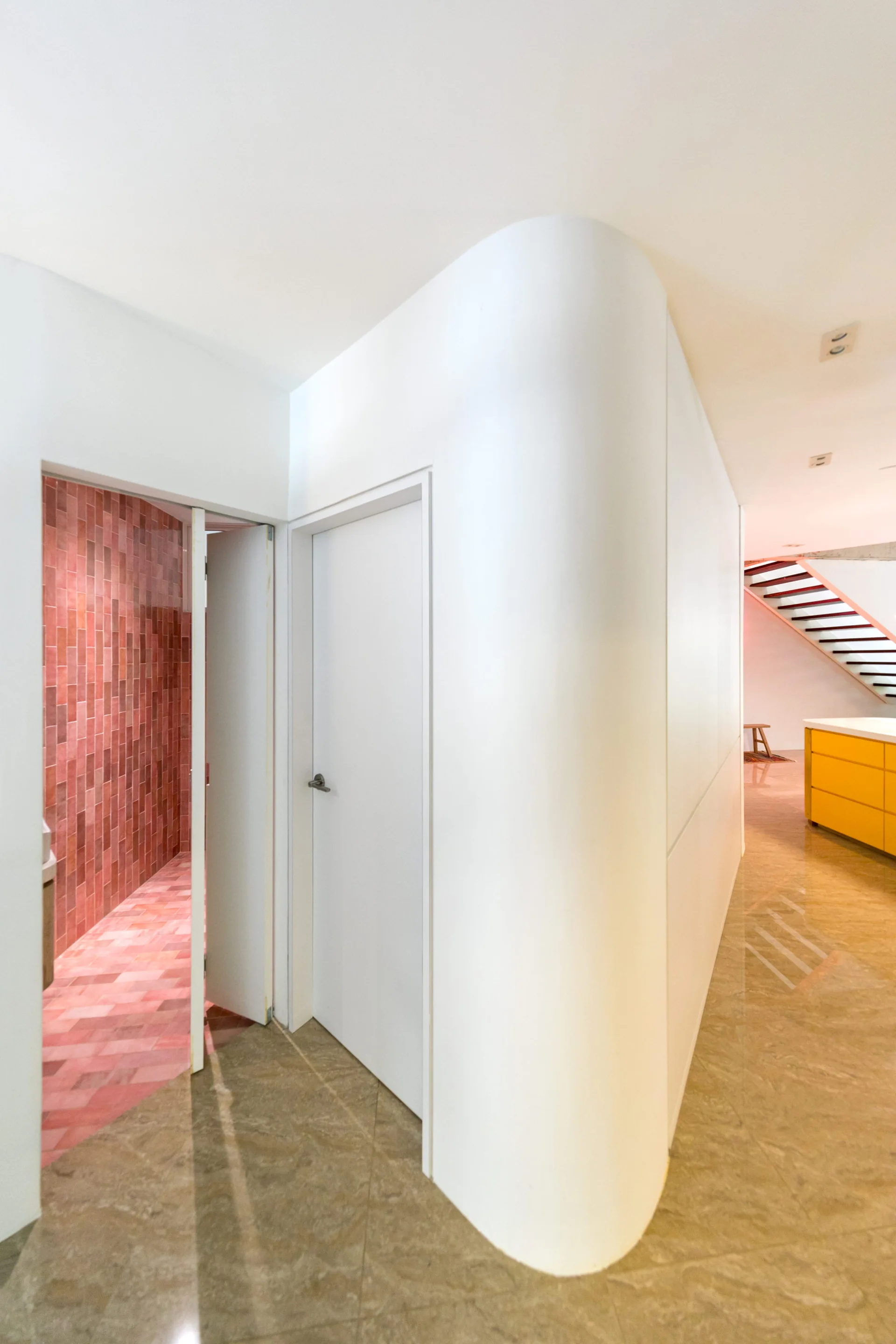 A modern white hallway features a prominent curved wall next to an open doorway revealing a bathroom covered in vertical red tiles.