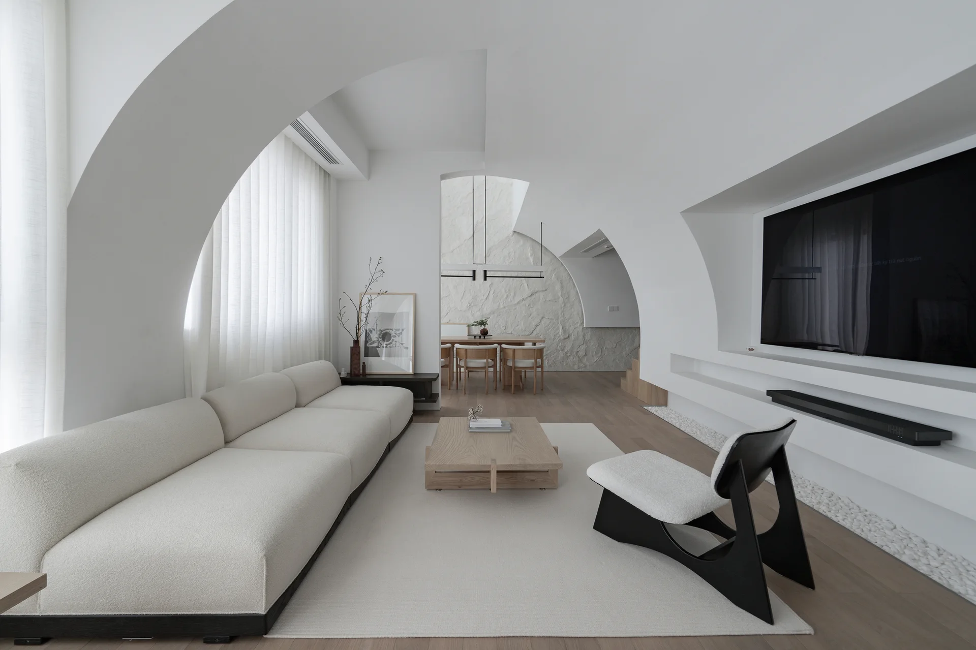 A minimalist living room featuring sculpted white plaster archways, a long cream sofa, a light wood coffee table, and a black accent chair.